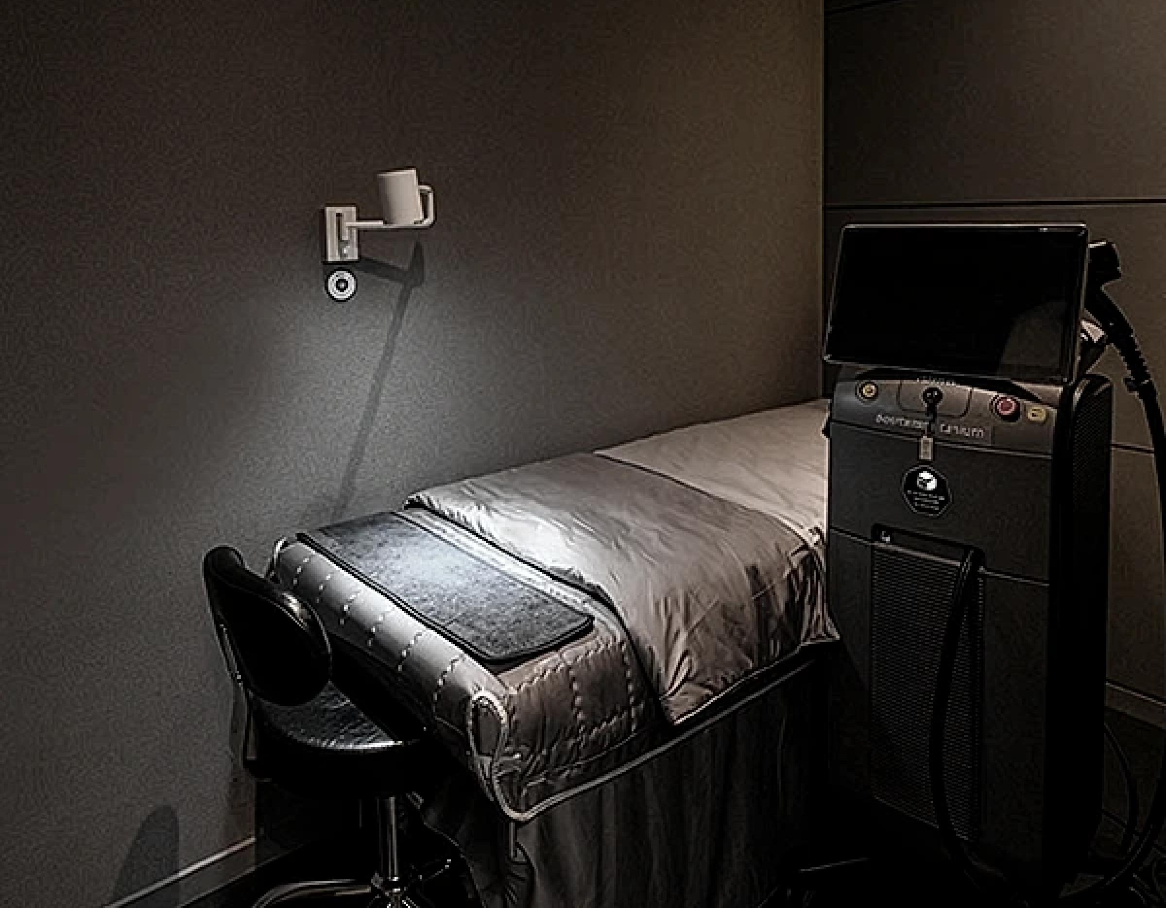 Private treatment room with a reclining bed, bedside lamp, black stool and medical laser device on a dark wall background