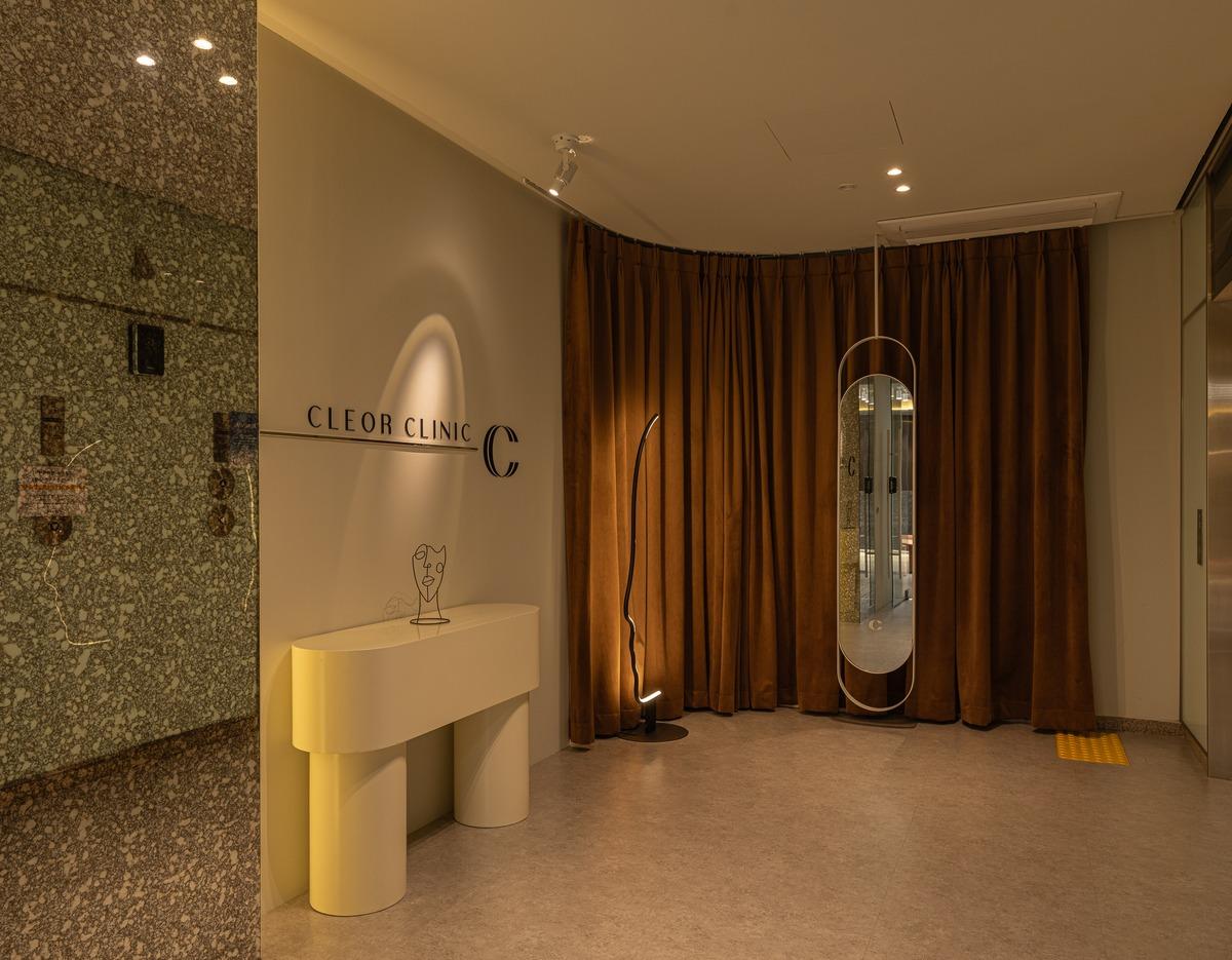 CLEOR Clinic Hongdae reception niche with beige console table, sculptural wire face decor, floor lamp, tall oval mirror and brown curtains