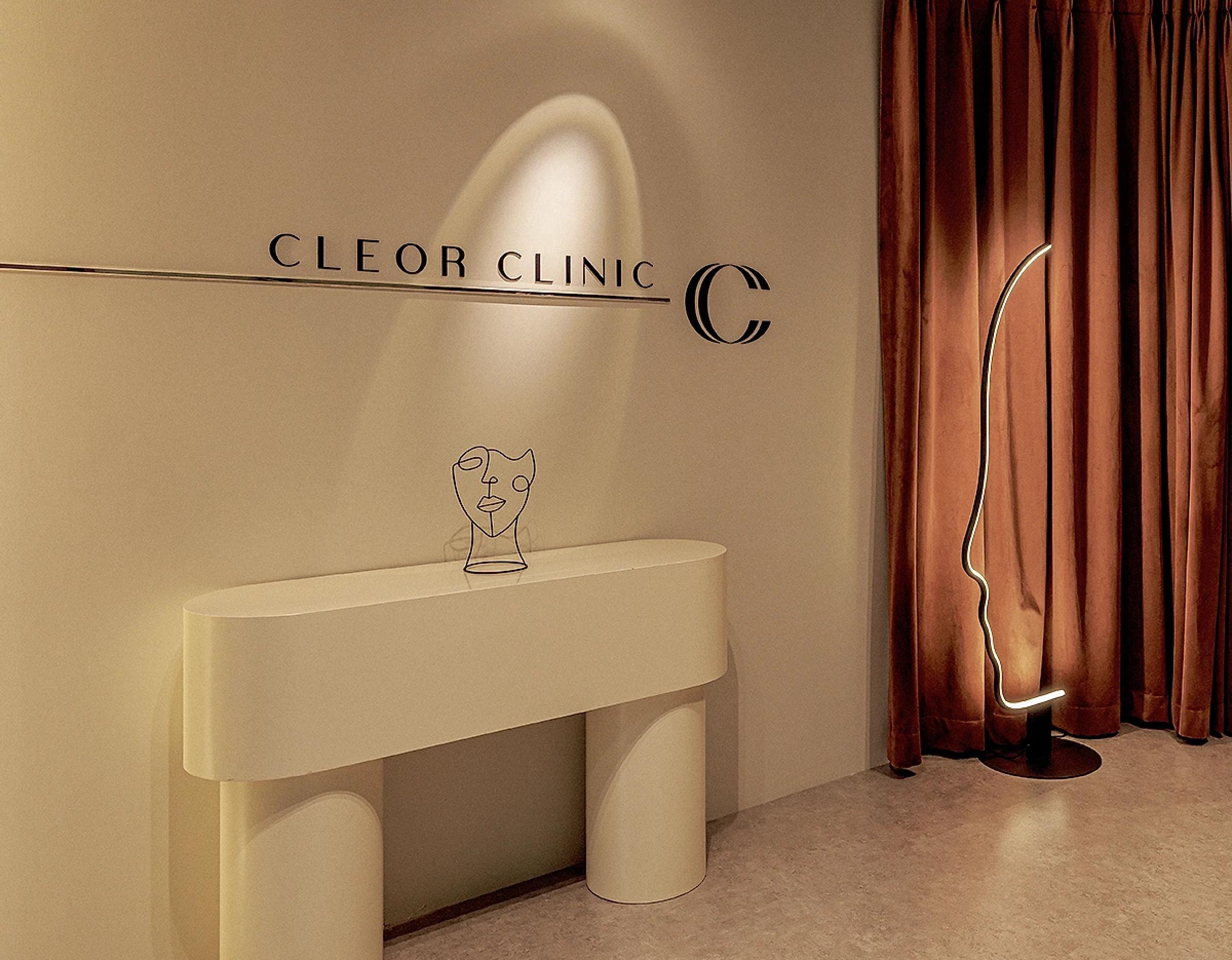 CLEOR CLINIC logo on beige wall above minimalist cream console table and sculptural face-shaped floor lamp by rust curtains