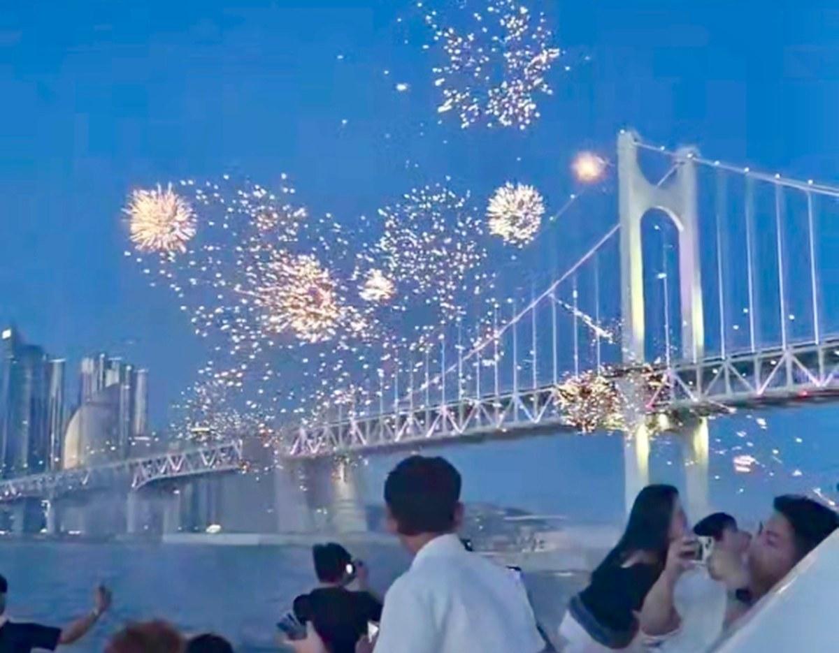 fireworks exploding over Gwangan Bridge at dusk with onlookers on a yacht photographing the display