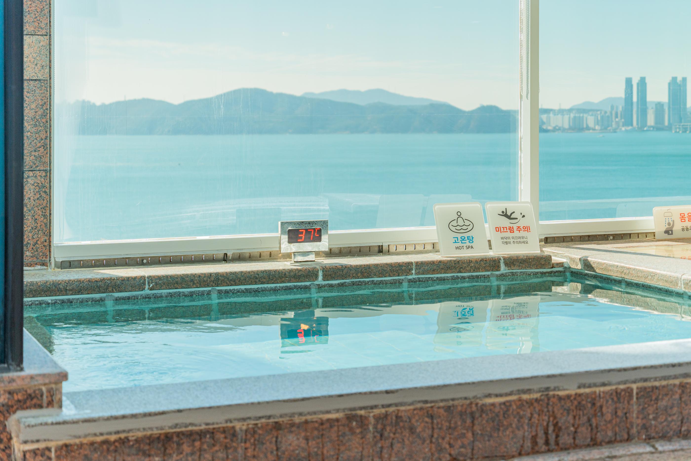 B-BEAUTY hot spa pool by a window with ocean and distant skyline, digital thermometer reading 37°C and safety signs visible