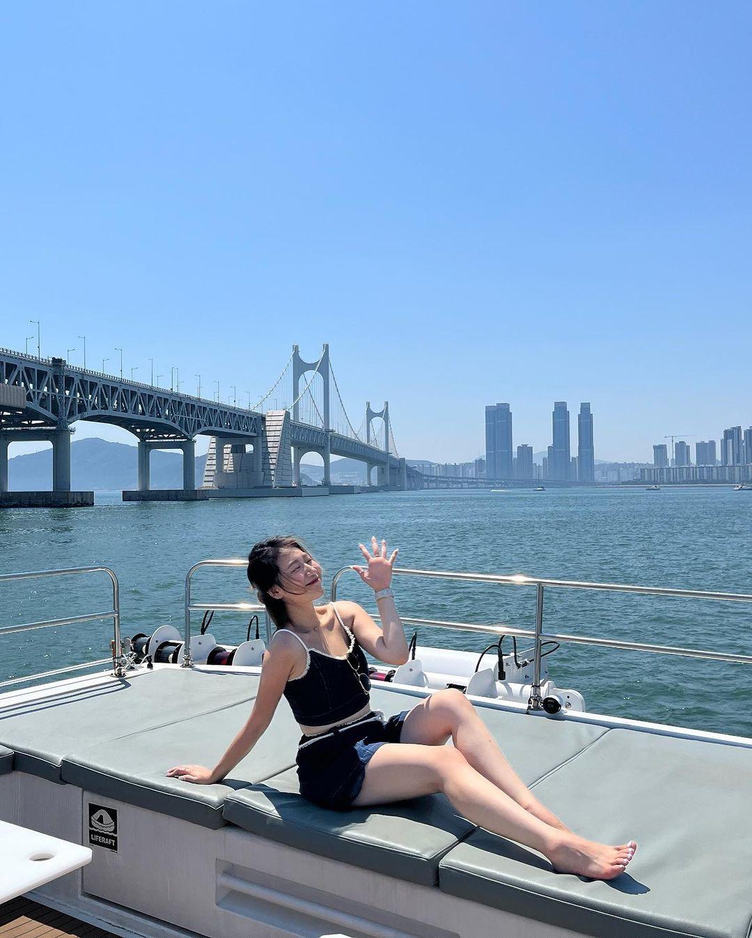 B-BEAUTY guest in black outfit lounging on yacht deck, waving with Gwangan Bridge and Busan skyline behind her