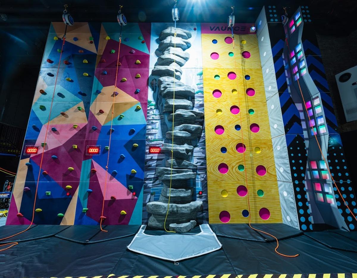 Indoor climbing wall area with colorful geometric panels, faux rock column, holey yellow wall and safety mats at Bounce Park 제주센터