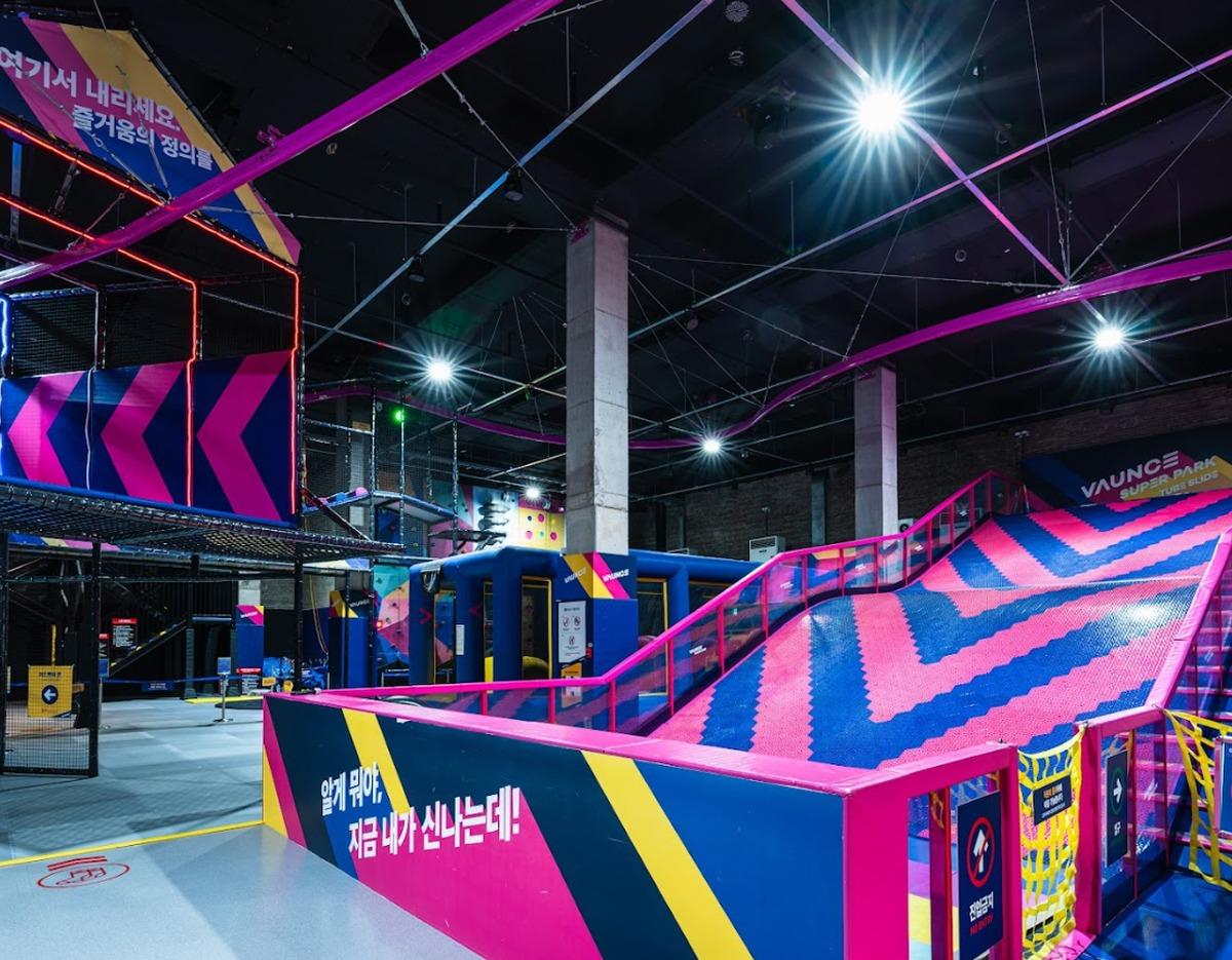 Indoor trampoline park with bright pink and blue foam slide, ramps, netted climbing structures and neon lighting.