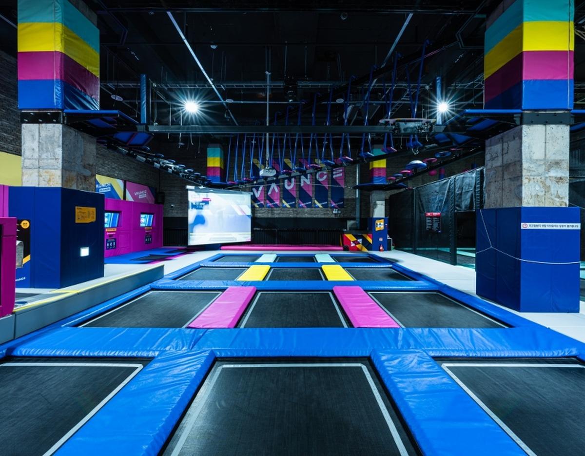 Indoor trampoline court with colorful padded frames, multiple rectangular jump beds and hanging aerial challenge elements above.