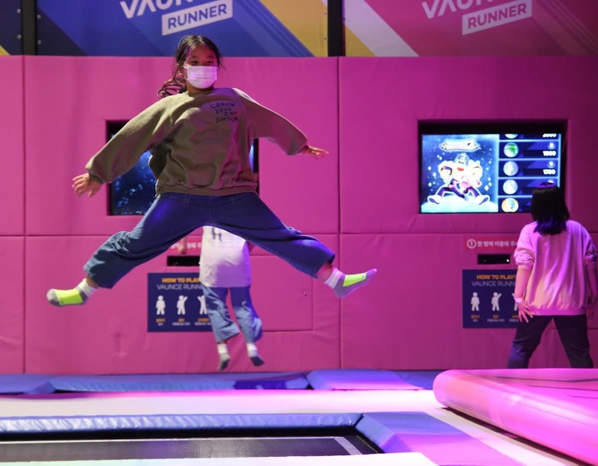 Young person wearing a mask and neon socks mid-split jump on a pink trampoline at 제주 바운스 슈퍼파크 제주센터.