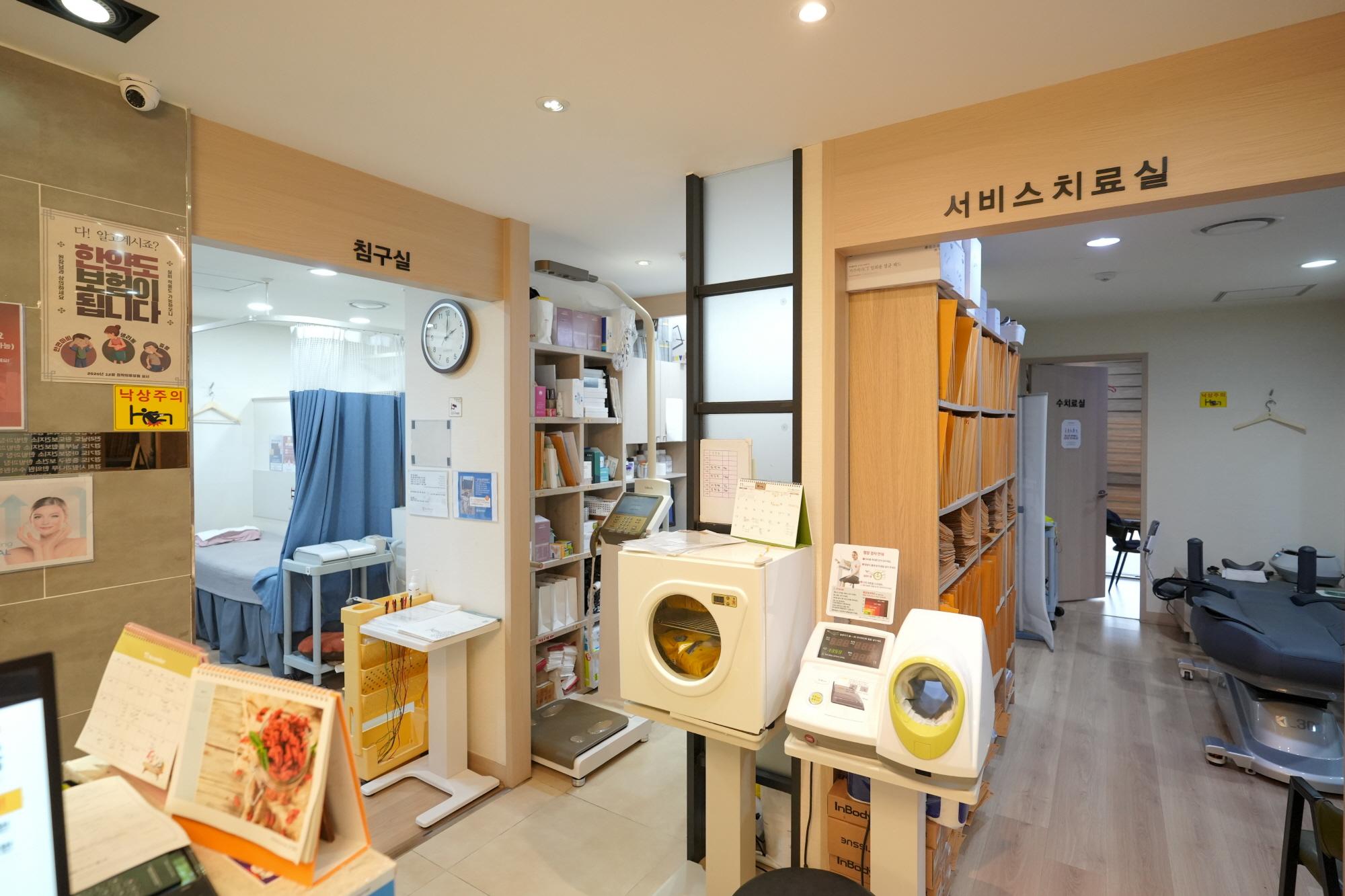 경희일생한의원 reception area with treatment rooms, shelving, medical devices like ultrasound/diagnostic machines and curtained acupuncture bed visible.