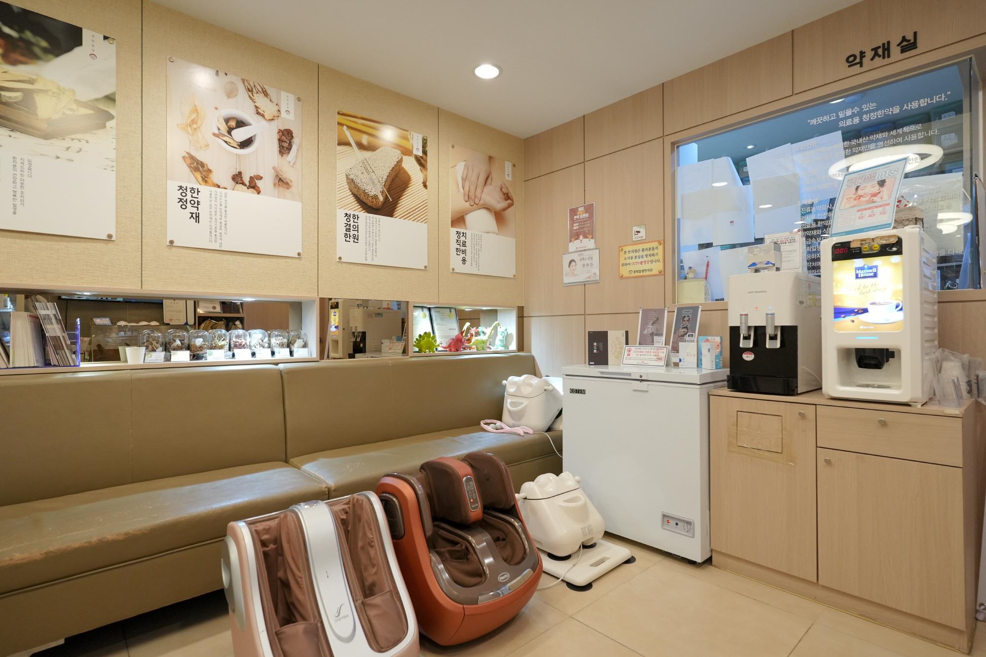 경희일생한의원 reception area with beige bench, foot and leg massage machines, water and coffee dispensers, and herbal posters on wall