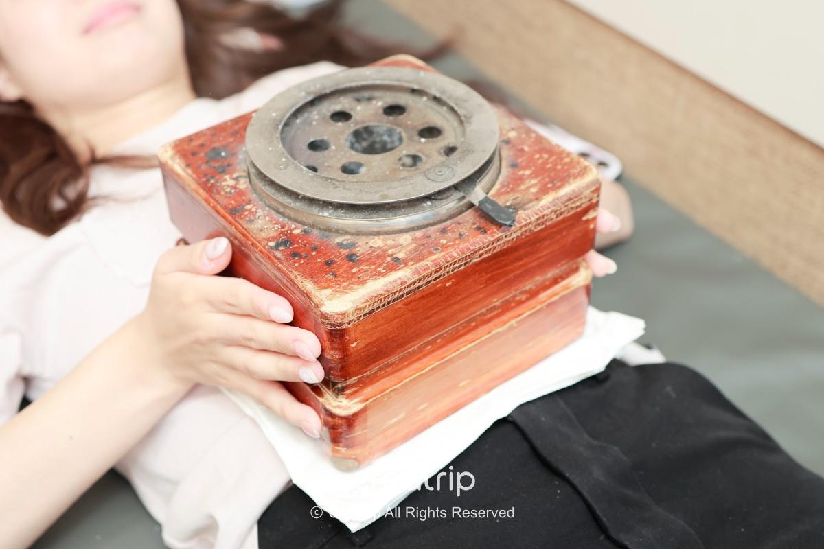 patient lying on back holding a stacked wooden moxibustion box with metal top placed on abdomen
