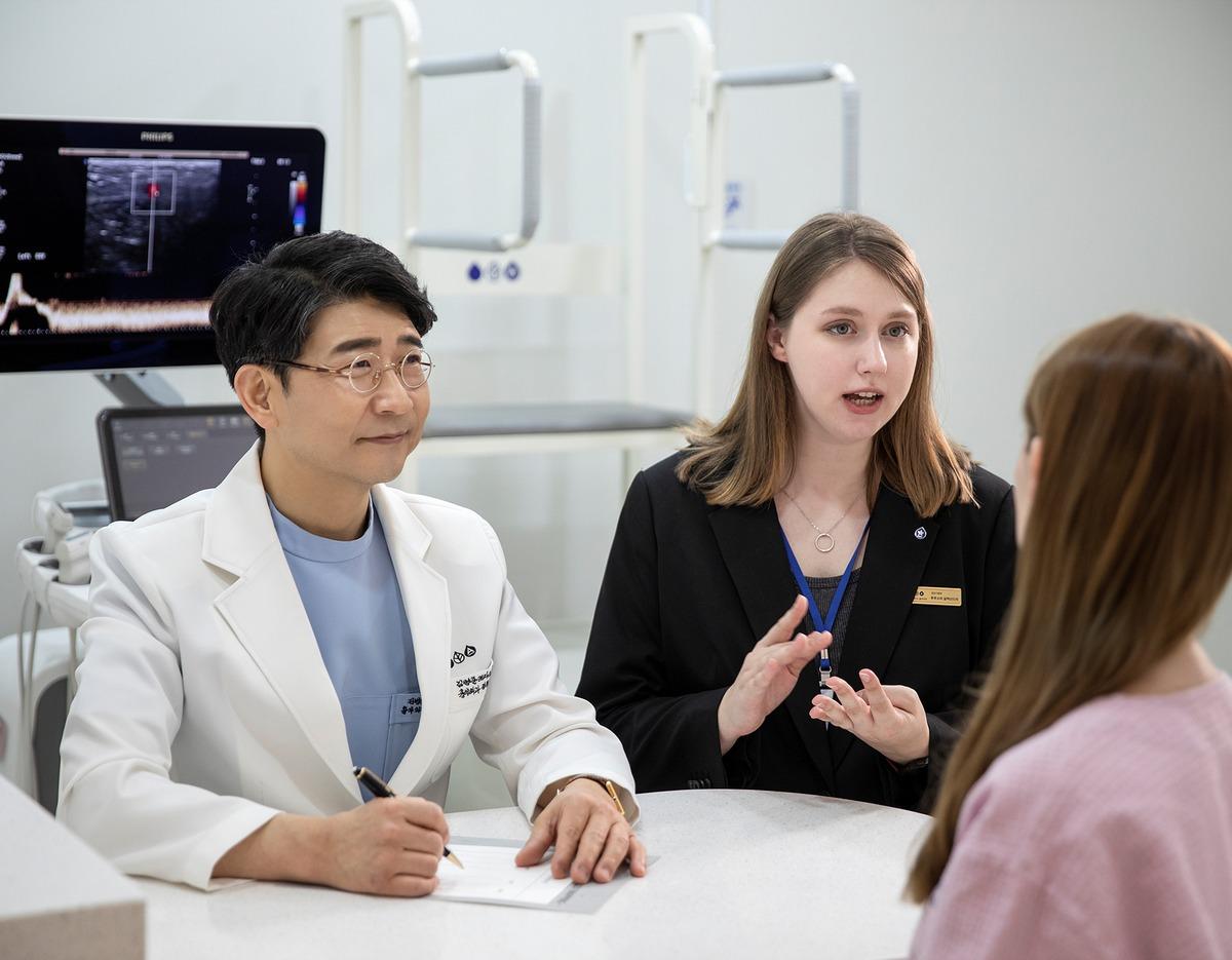 Doctor in white coat writing on a form while a staff member in a black blazer explains treatment to a seated patient.
