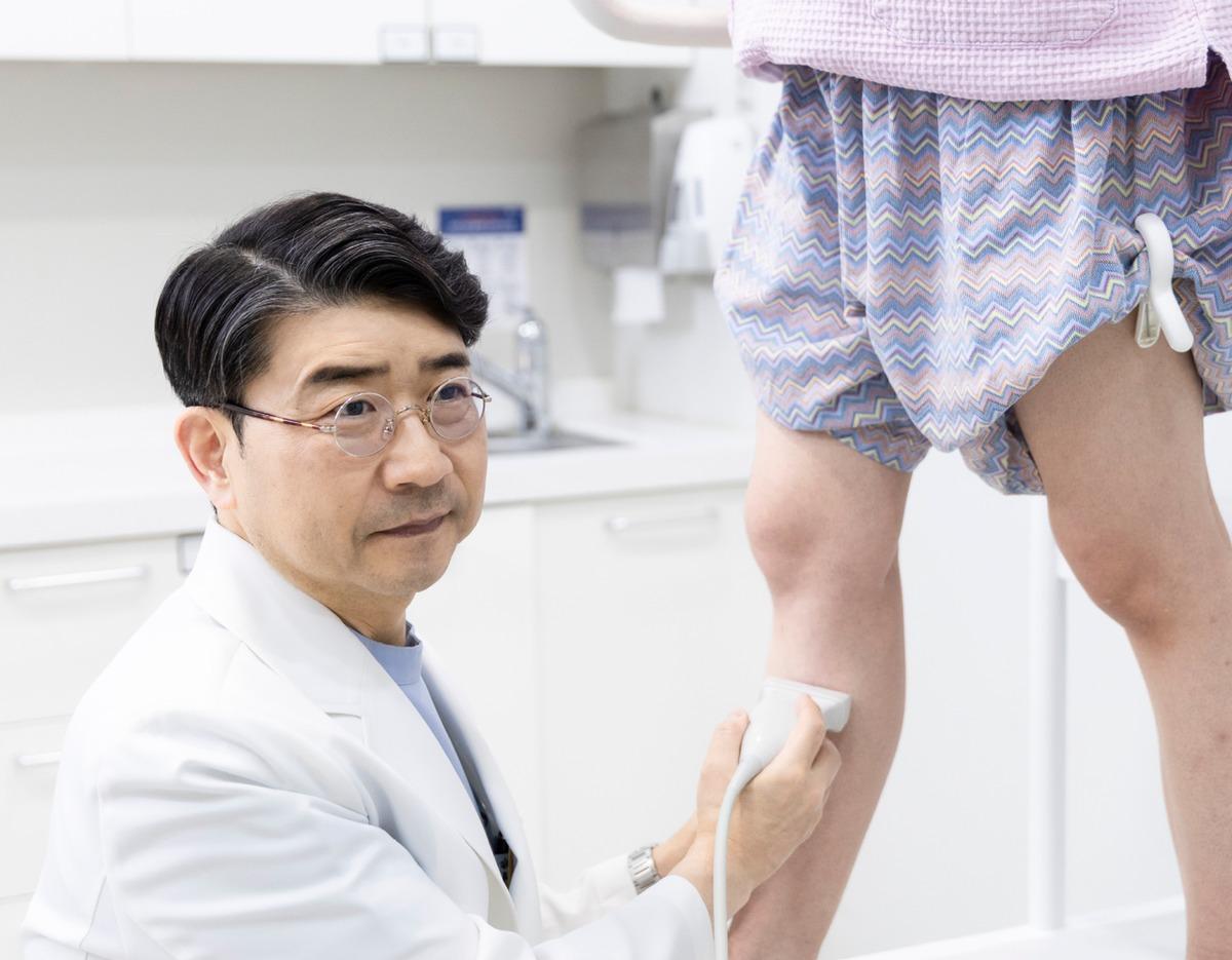 Doctor in white coat using an ultrasound probe on a patient's lower leg in a clinical exam room