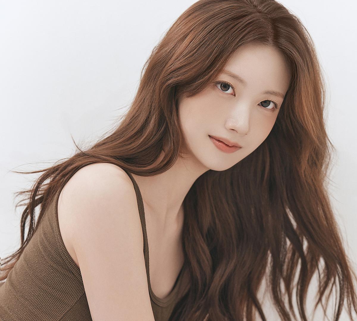 young woman with long brown hair and natural makeup wearing a brown tank top against a white backdrop