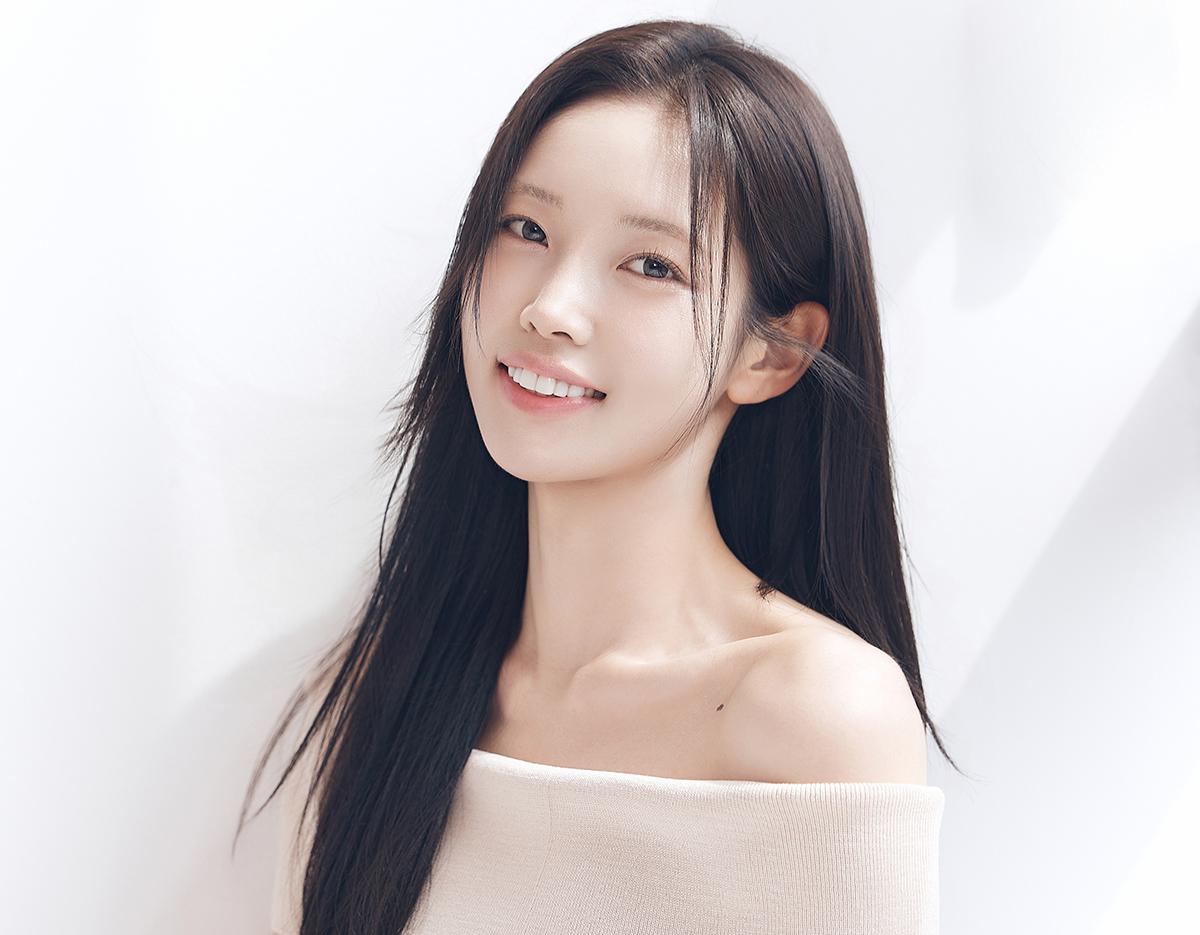 young woman with long dark hair and bare-shoulder beige top smiling against a soft white background