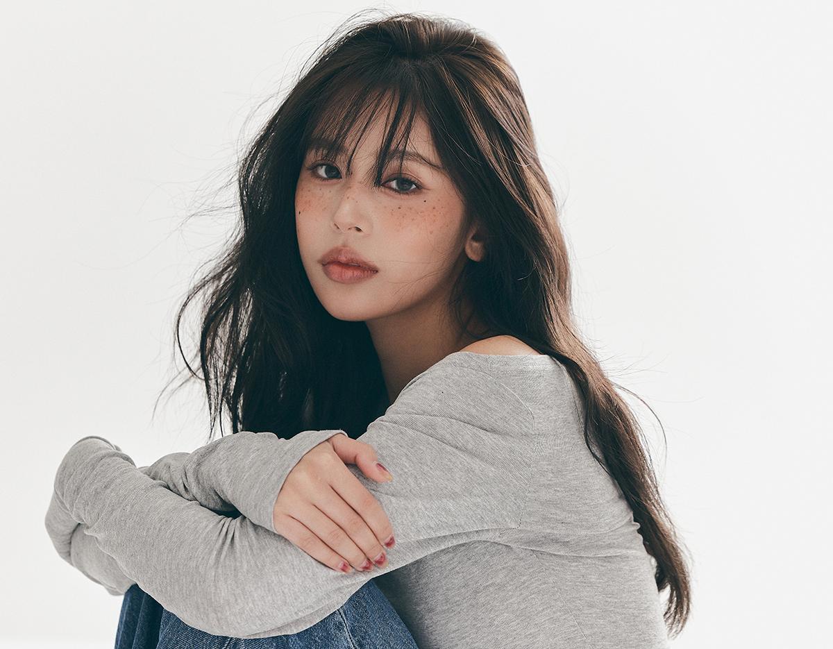young woman with long dark hair and freckles in gray top and jeans seated hugging knees against plain white background