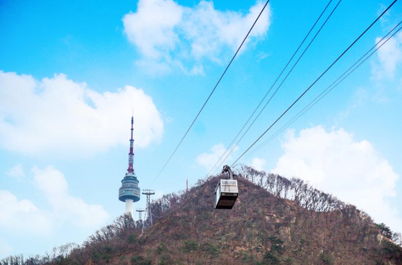 Namsan cable car cabin suspended on cables above a tree-covered hill with N Seoul Tower in the background, clear blue sky