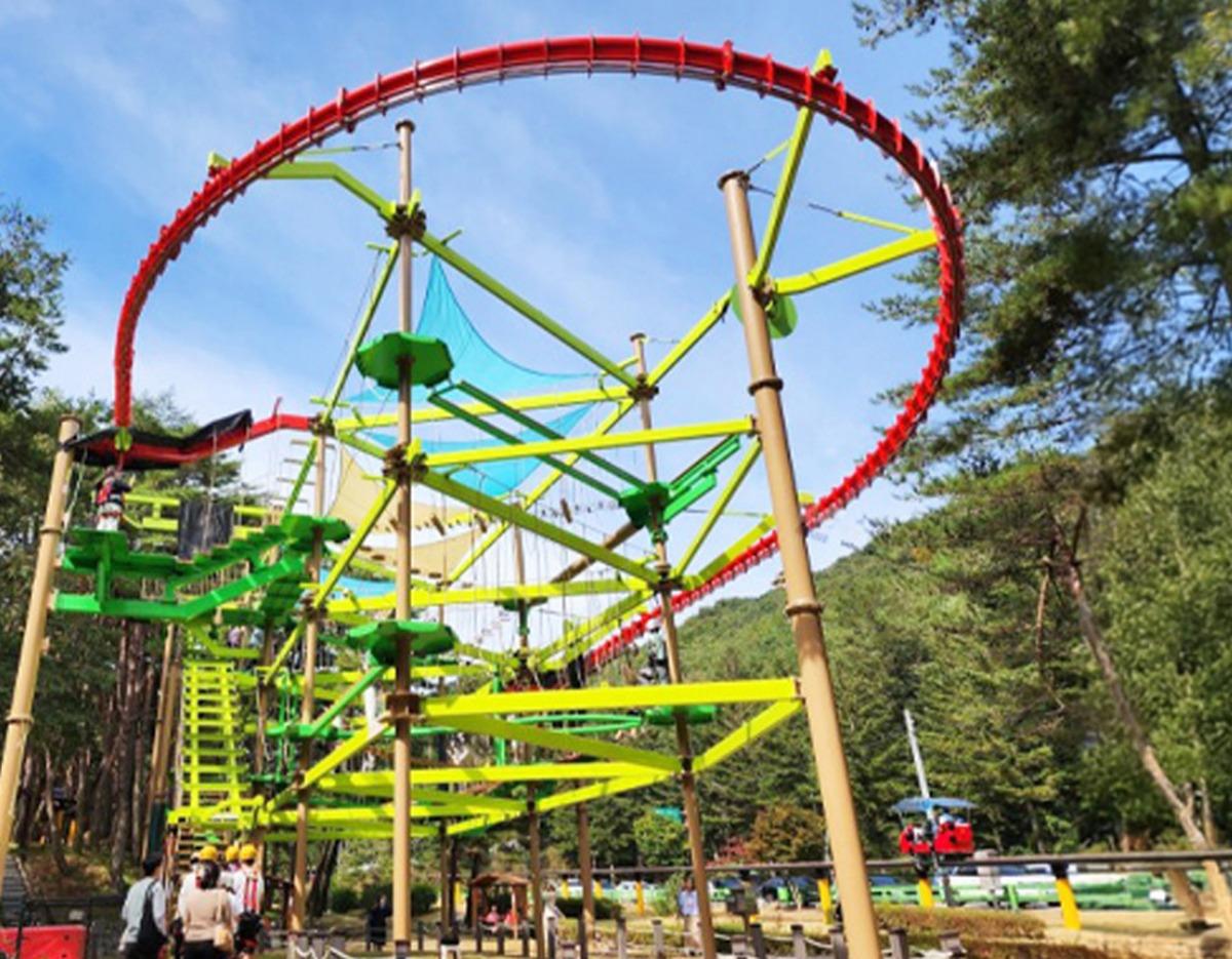 Outdoor multi-level green and yellow ropes course structure with red elevated circular track and people below at a forested attraction
