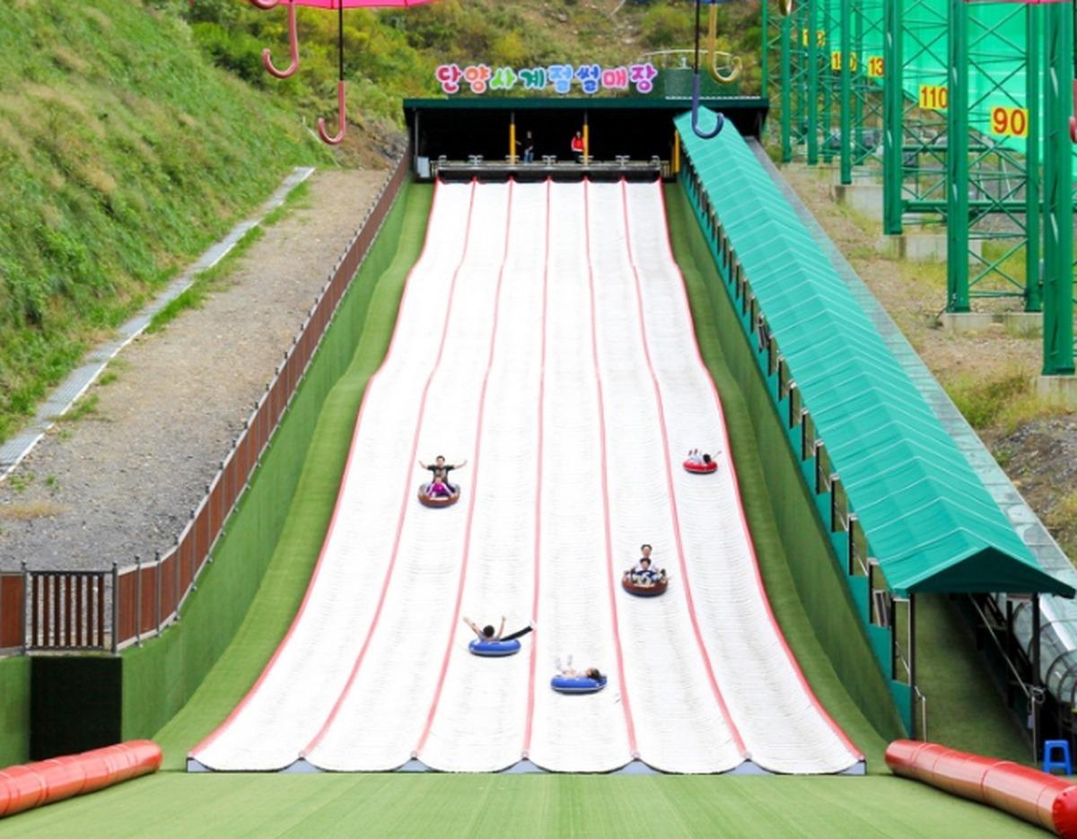 Outdoor multi-lane artificial snow tubing slope with riders on colorful inner tubes sliding down beside a covered conveyor lift