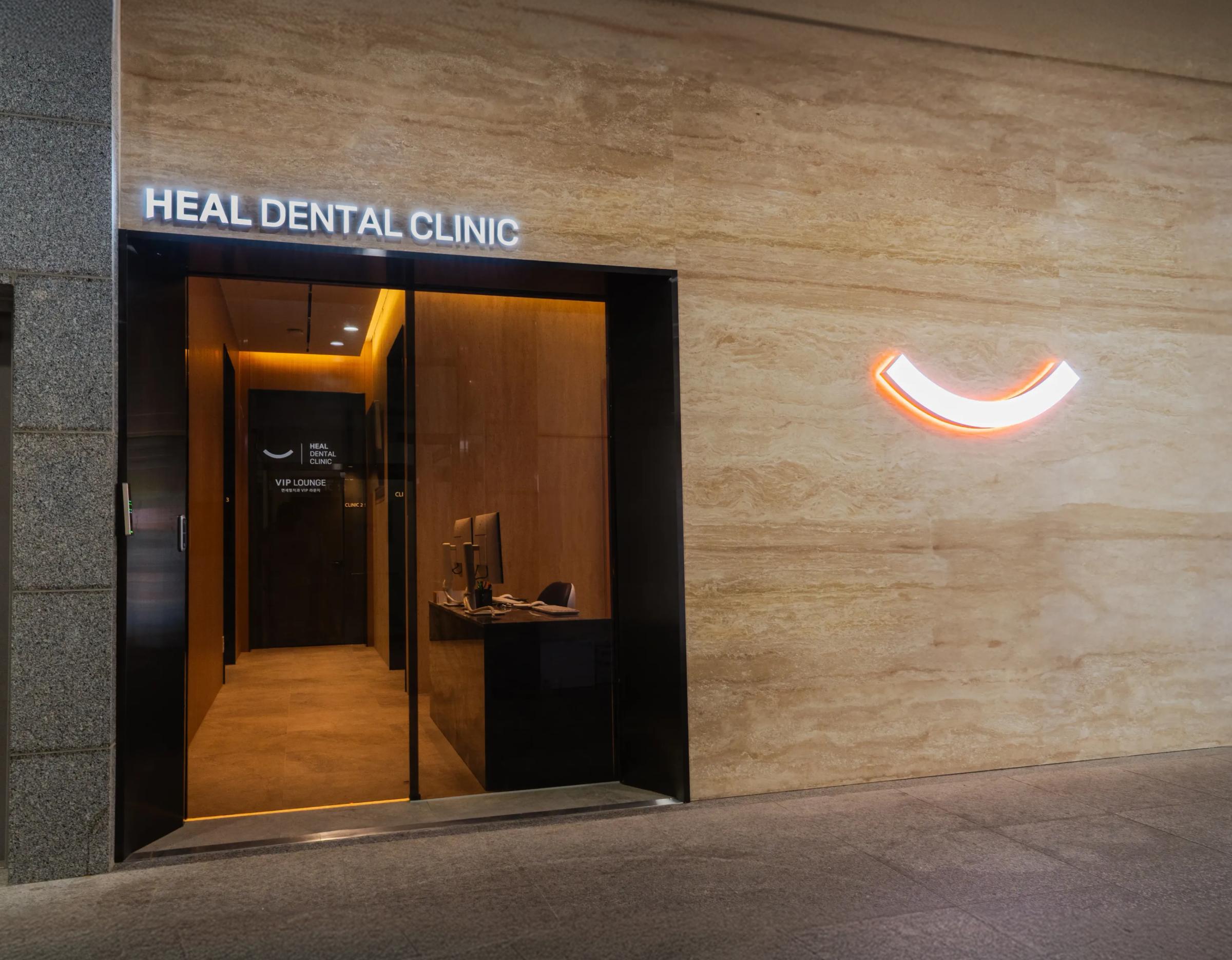 Yonsei Heal Dental Clinic entrance with illuminated "HEAL DENTAL CLINIC" sign, glass door, reception desk and smiling arc logo on wall