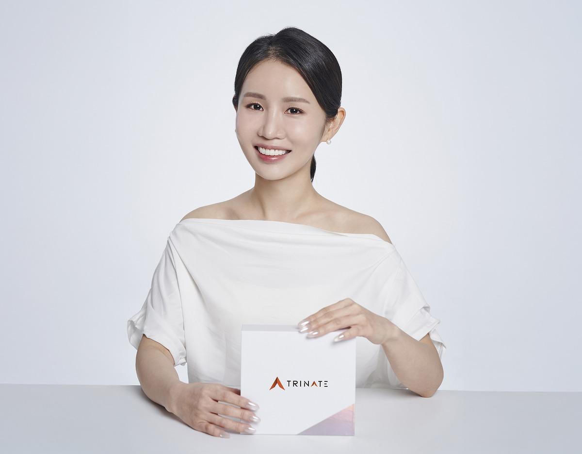 smiling woman in white top holding a Trinate product box on a white tabletop, clean studio portrait