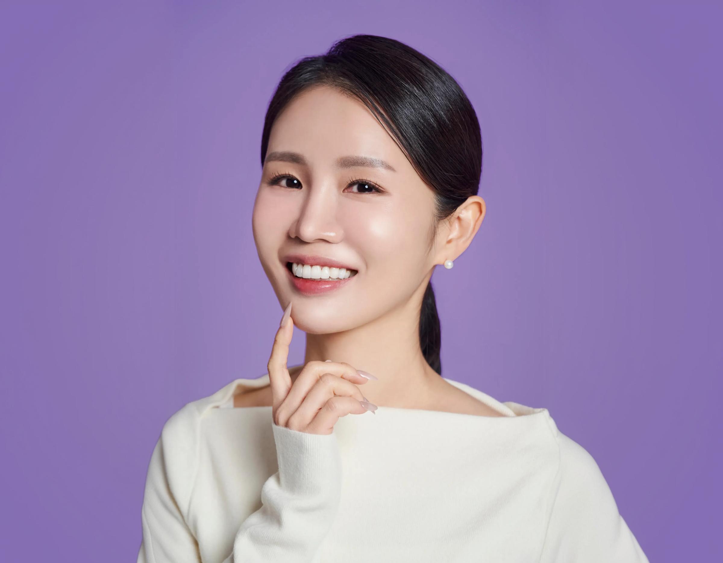 Smiling woman with pulled-back hair wearing white top, hand near chin, showing bright teeth against purple background