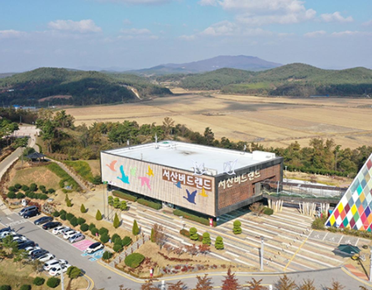 Seosan Birdland visitor center building with bird murals, parking lot, landscaped paths and surrounding fields and hills