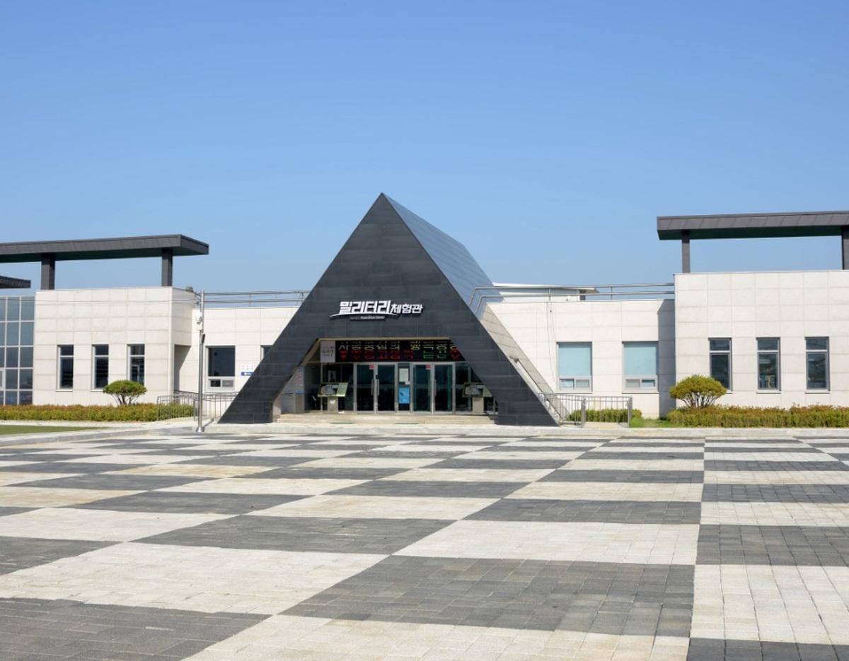 CHUNGNAM TOURPASS: modern low white building with a black triangular entrance and wide checkerboard plaza under clear blue sky