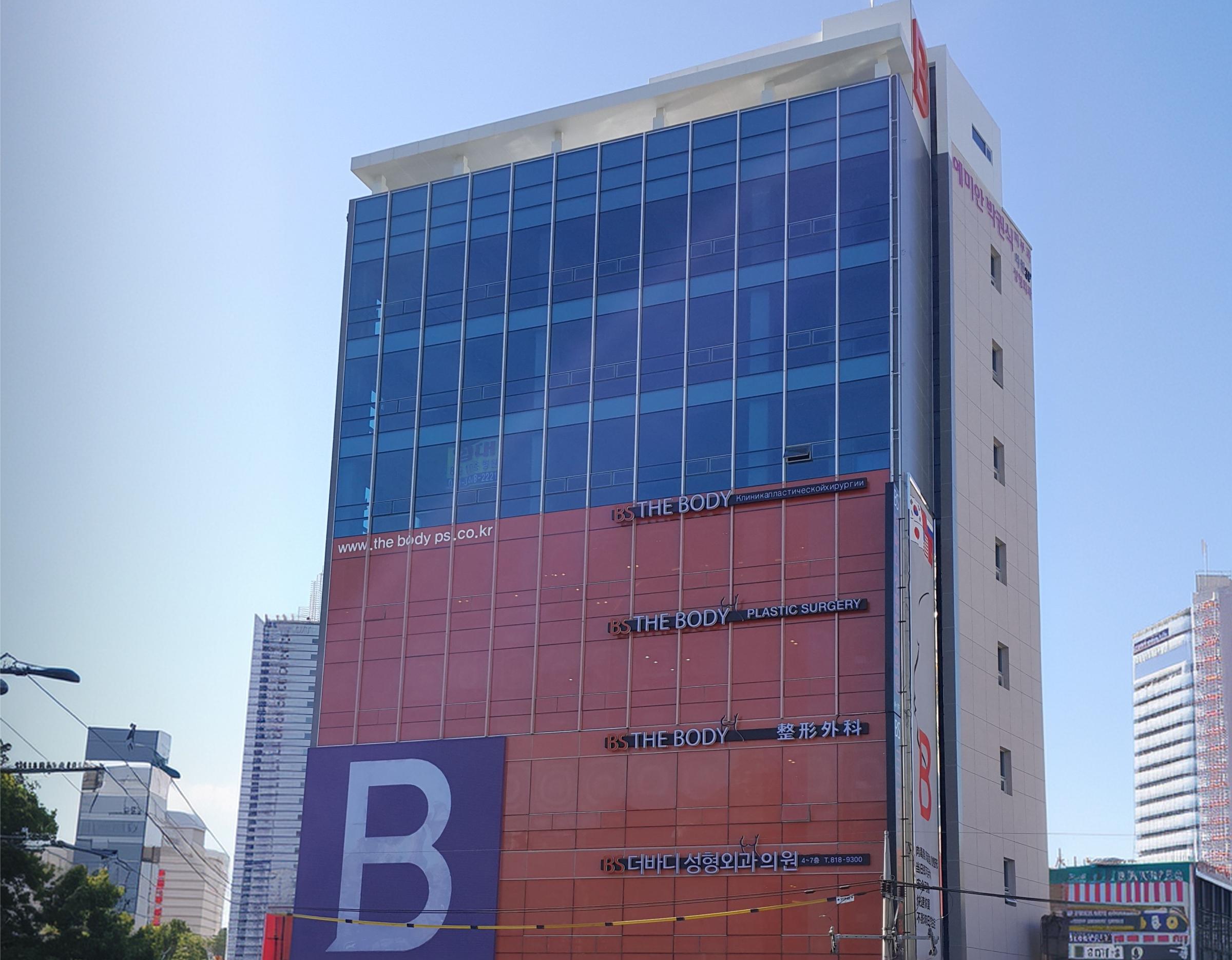 Exterior of 더바디성형외과의원 building in Busan Seomyeon, multi-story glass facade with large purple "B" sign.