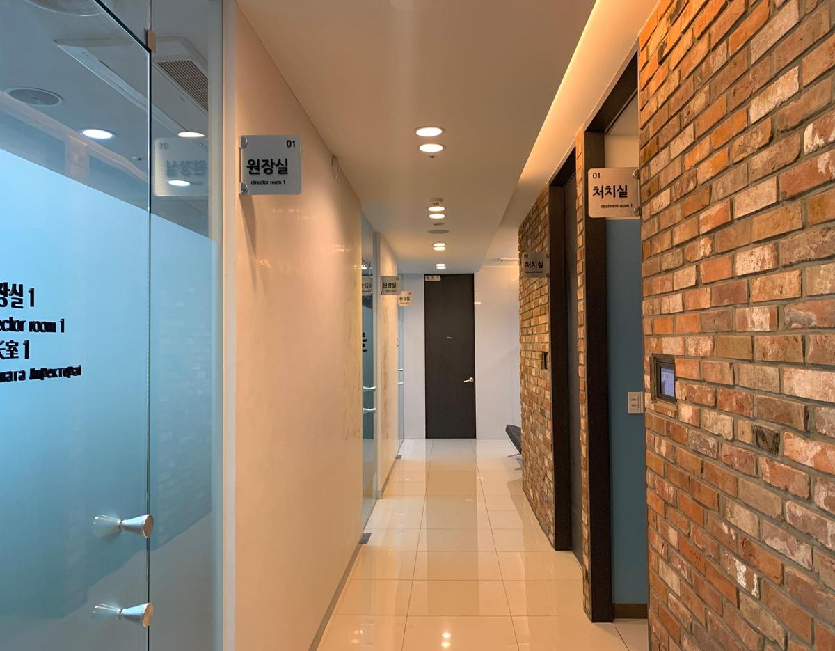 Clinic hallway with glossy tiled floor, glass doors on left and brick-faced treatment rooms on right at The Body (Busan Seomyeon).