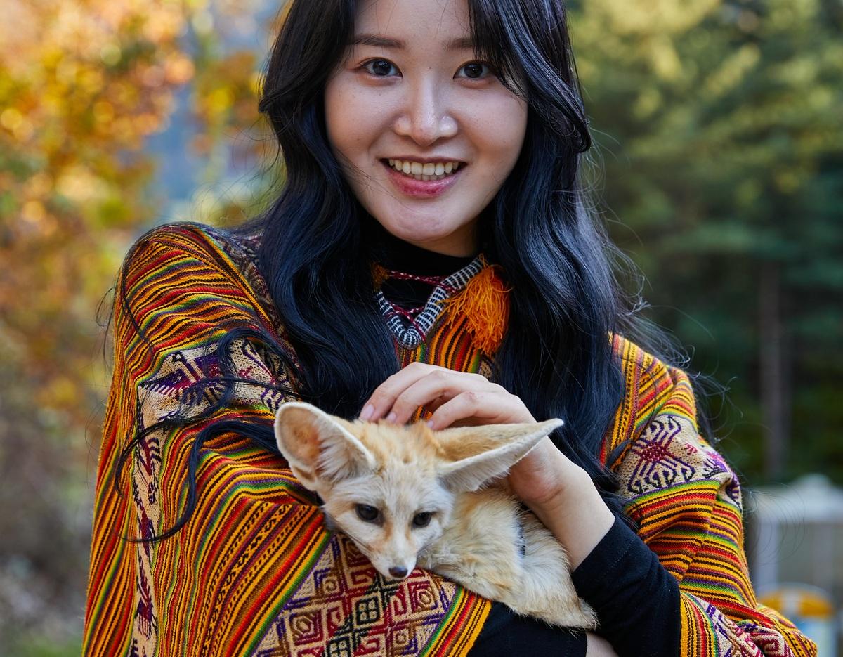 young woman in colorful striped jacket smiling while cradling a small fennec fox with large ears against an autumn forest background