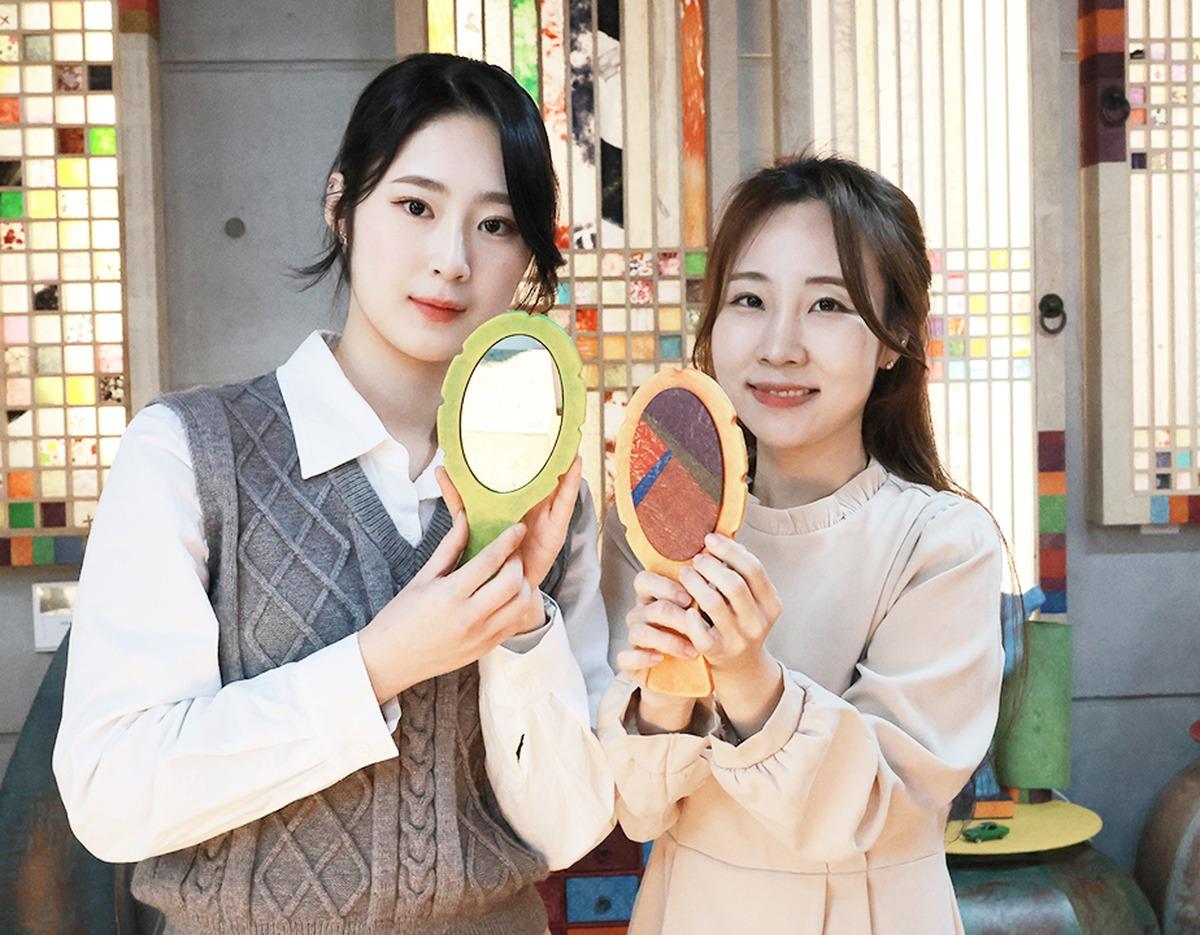 Two women in a craft studio holding handmade hanji hand mirrors with colorful paper insets, smiling in front of patterned doors