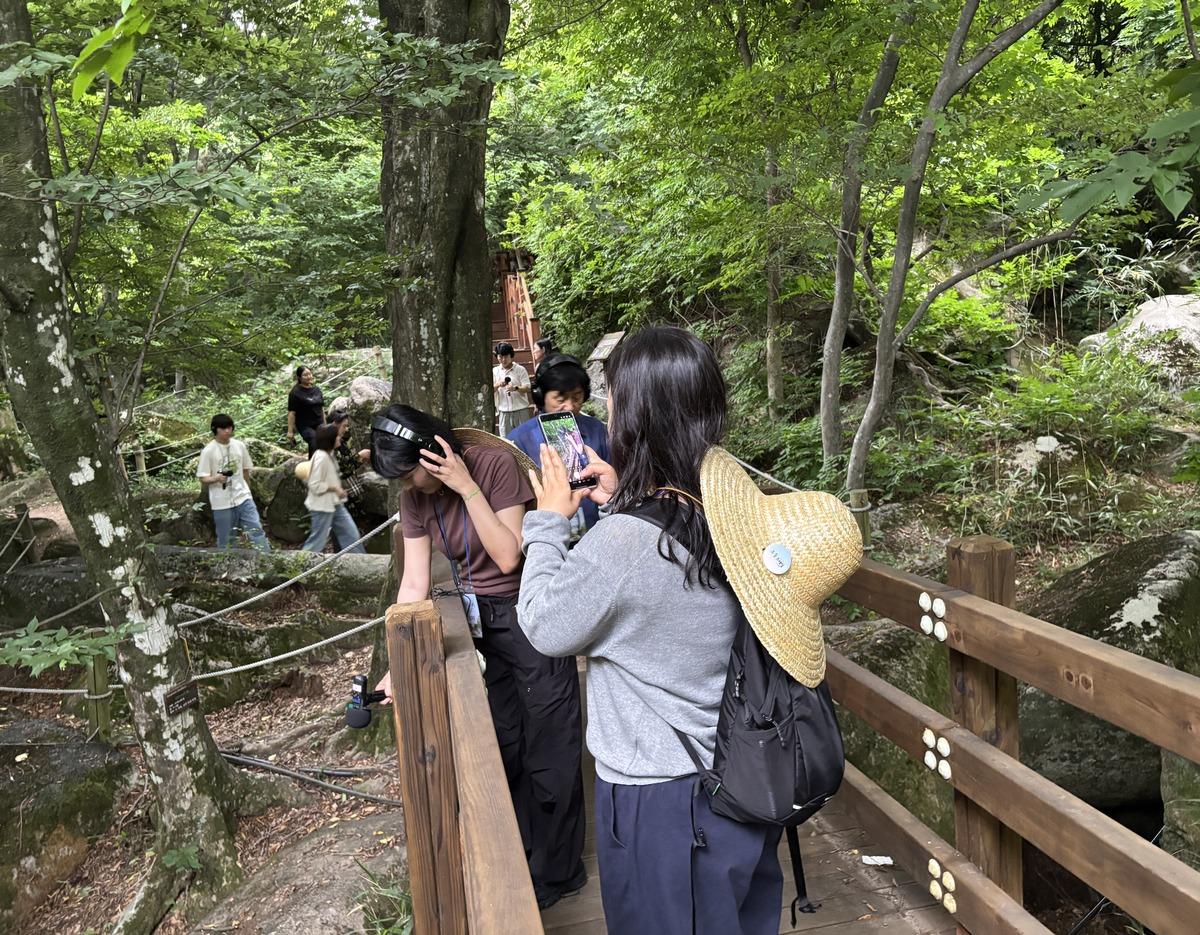 Group of hikers on a wooden forest trail in Geumjeongsan; woman in straw hat and backpack takes photo while others listen and explore.