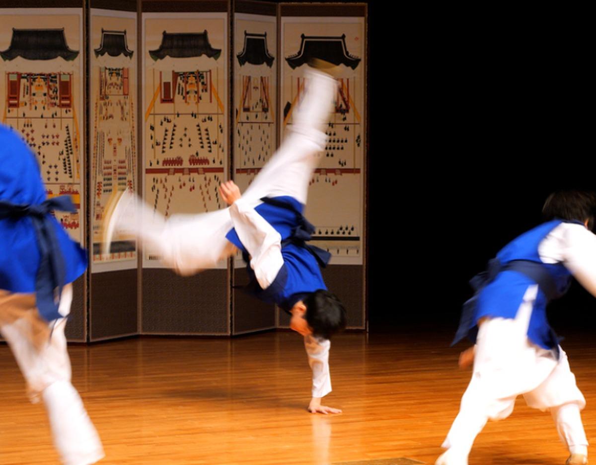 Young Korean traditional musician mid-air cartwheel on stage wearing blue hanbok-style performance outfit at Seoul Namsan Gugakdang.