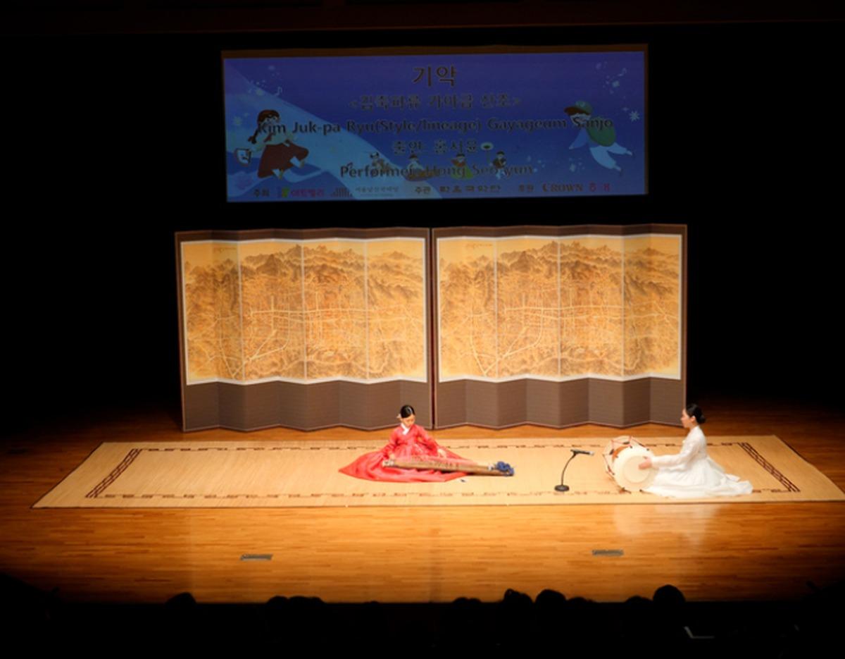 Seated young gayageum player in red and drummer in white performing on stage in front of a folding landscape screen