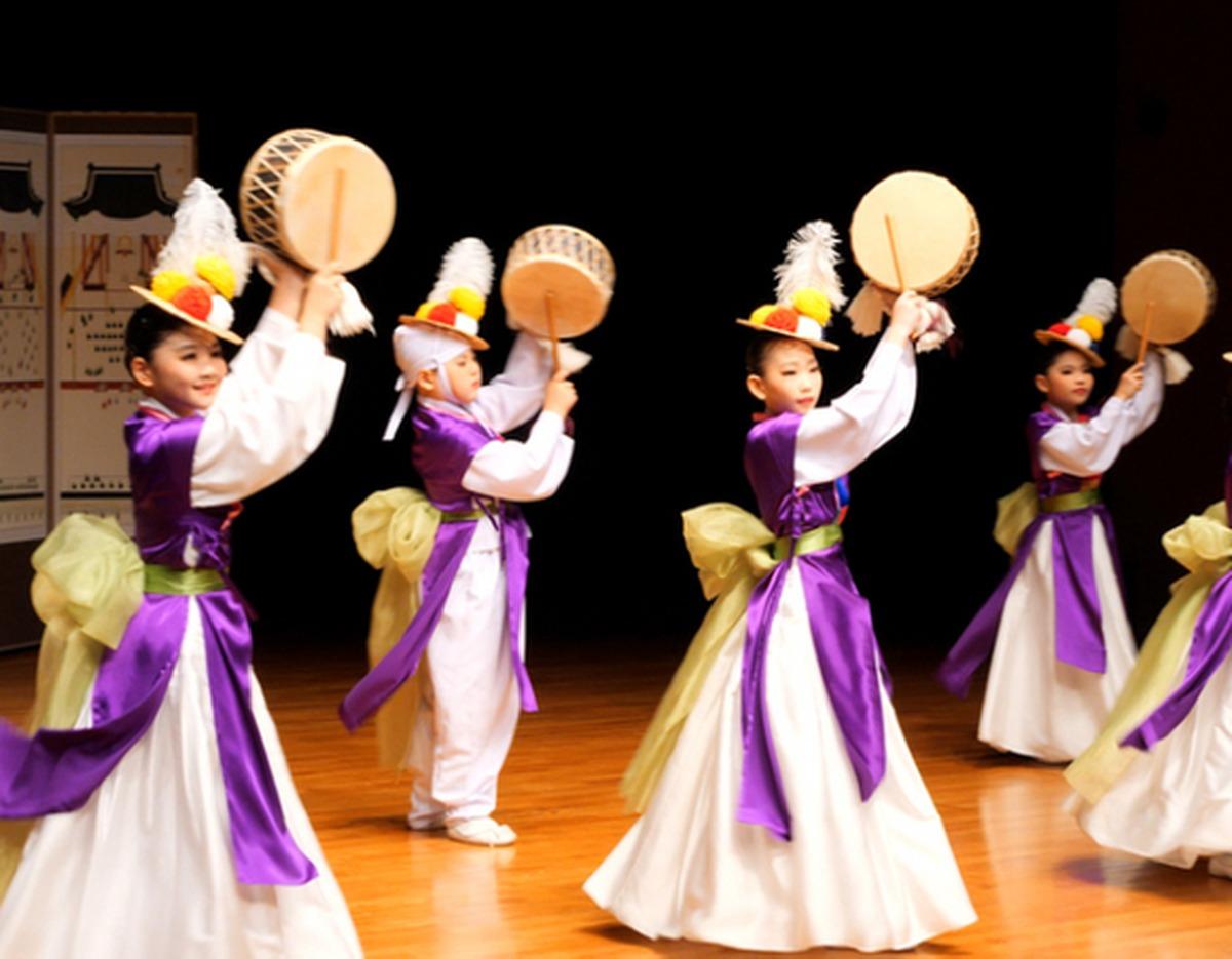 Young Korean performers in purple and white hanbok with feathered hats playing handheld drums on stage at Seoul Namsan Gugakdang