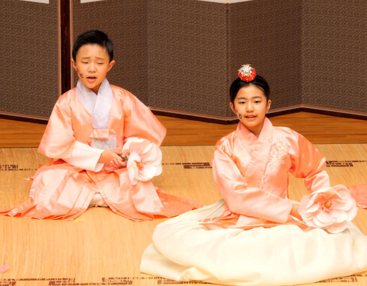 Two young performers in peach hanbok seated on a wooden stage holding large pink lotus props during a traditional music performance