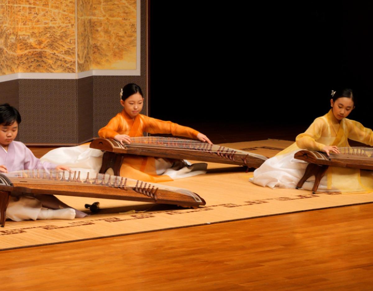 Three young Korean musicians in colorful hanbok seated on stage playing gayageum zithers during a traditional music performance