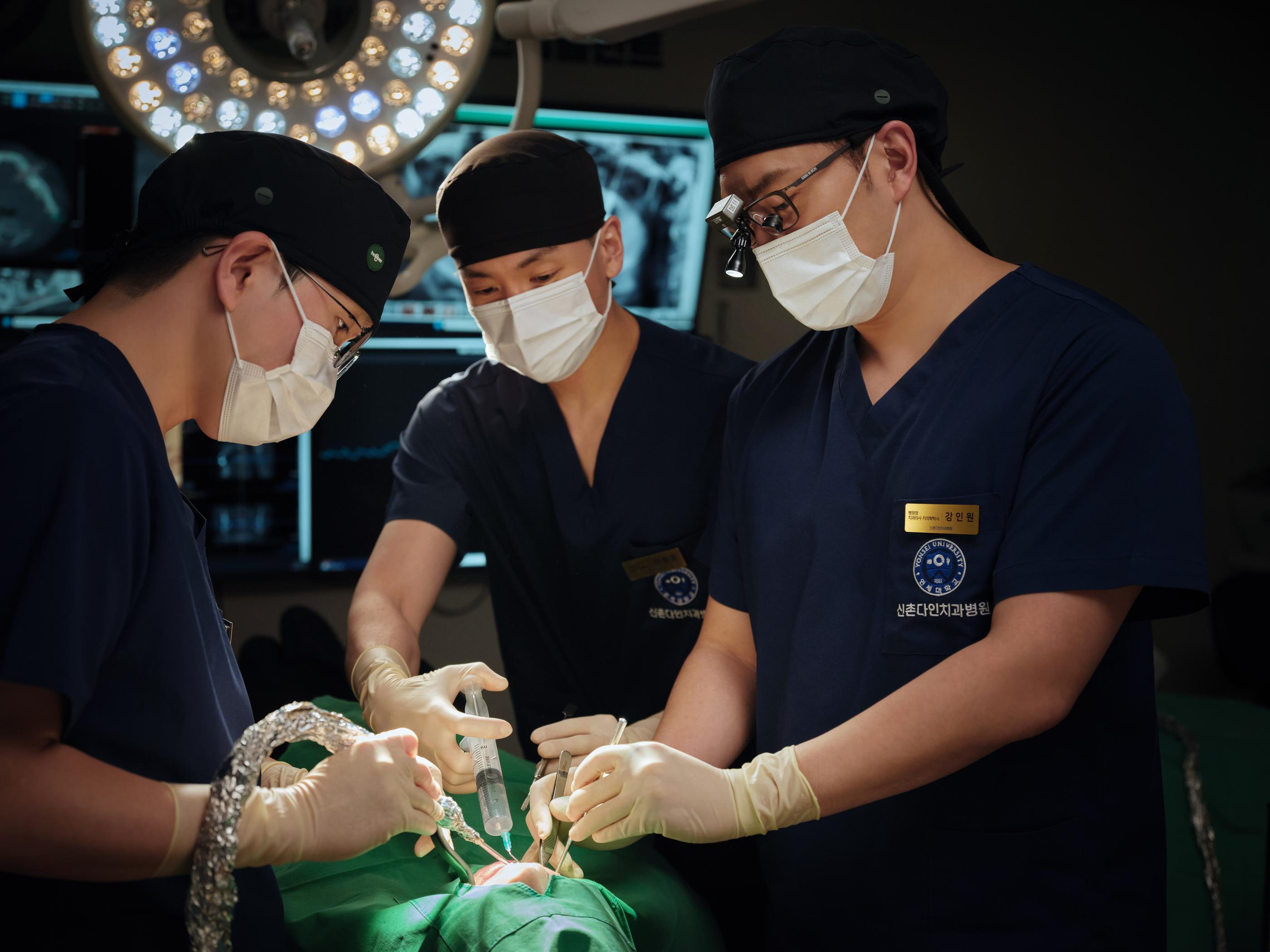 Surgical team at 신촌다인치과병원 in navy scrubs and masks performing dental operation under surgical light with instruments and syringe.