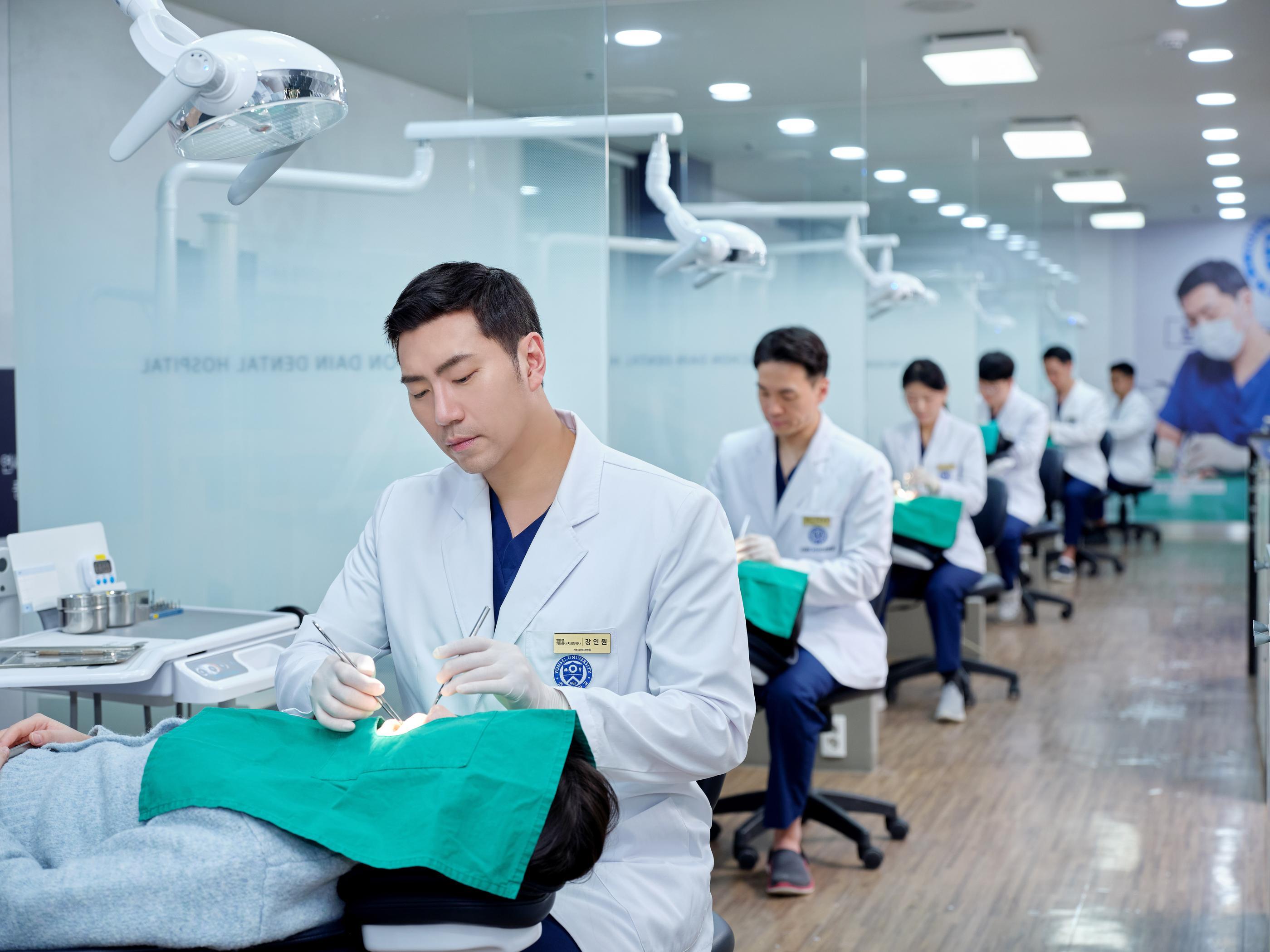 Sinchon Dain Dental Hospital dentist in white coat treating reclining patient under dental lamp, row of dentists in background.