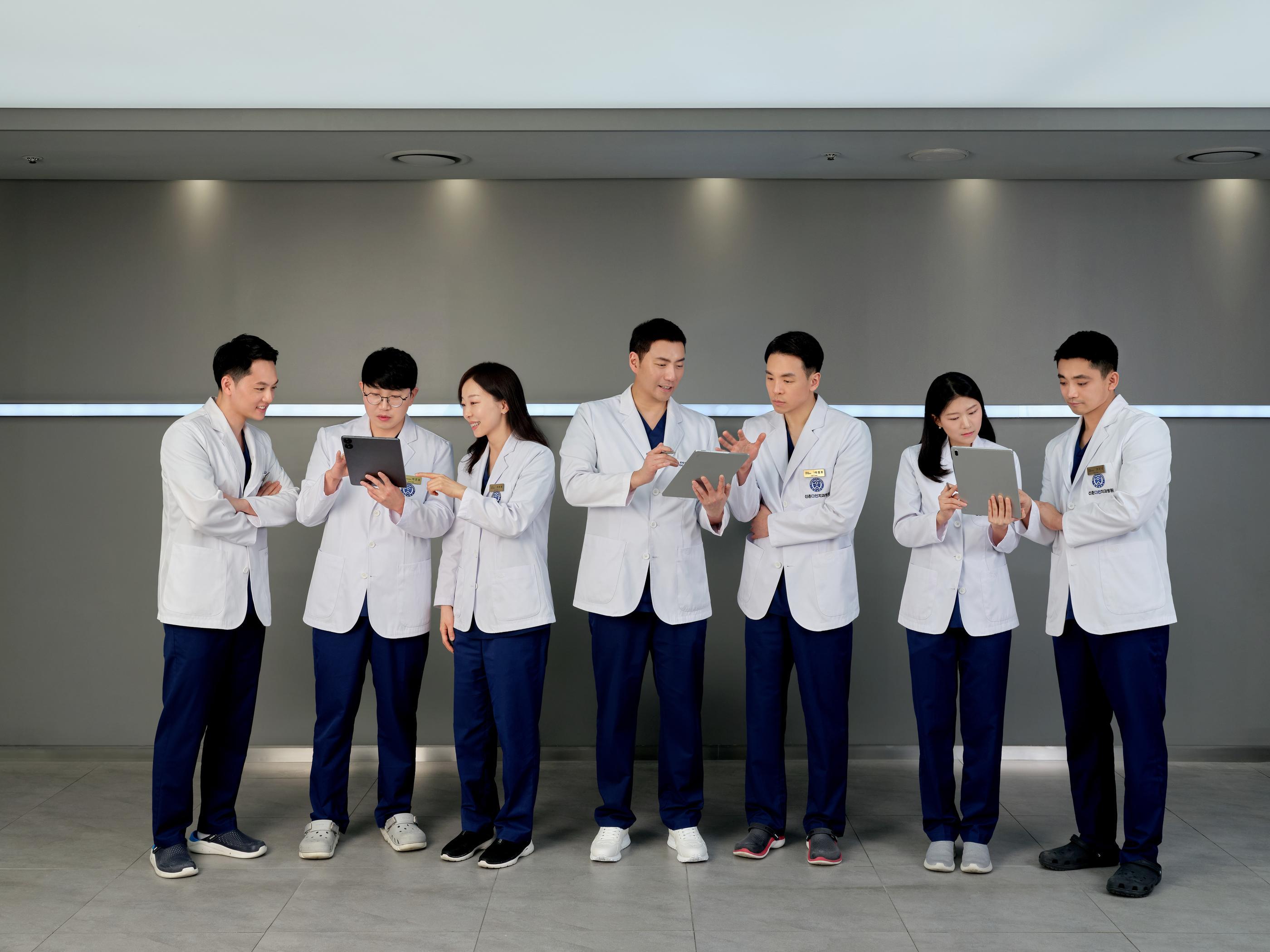 Sinchon Dain Dental Hospital medical team of seven in white coats reviewing tablets together against grey wall