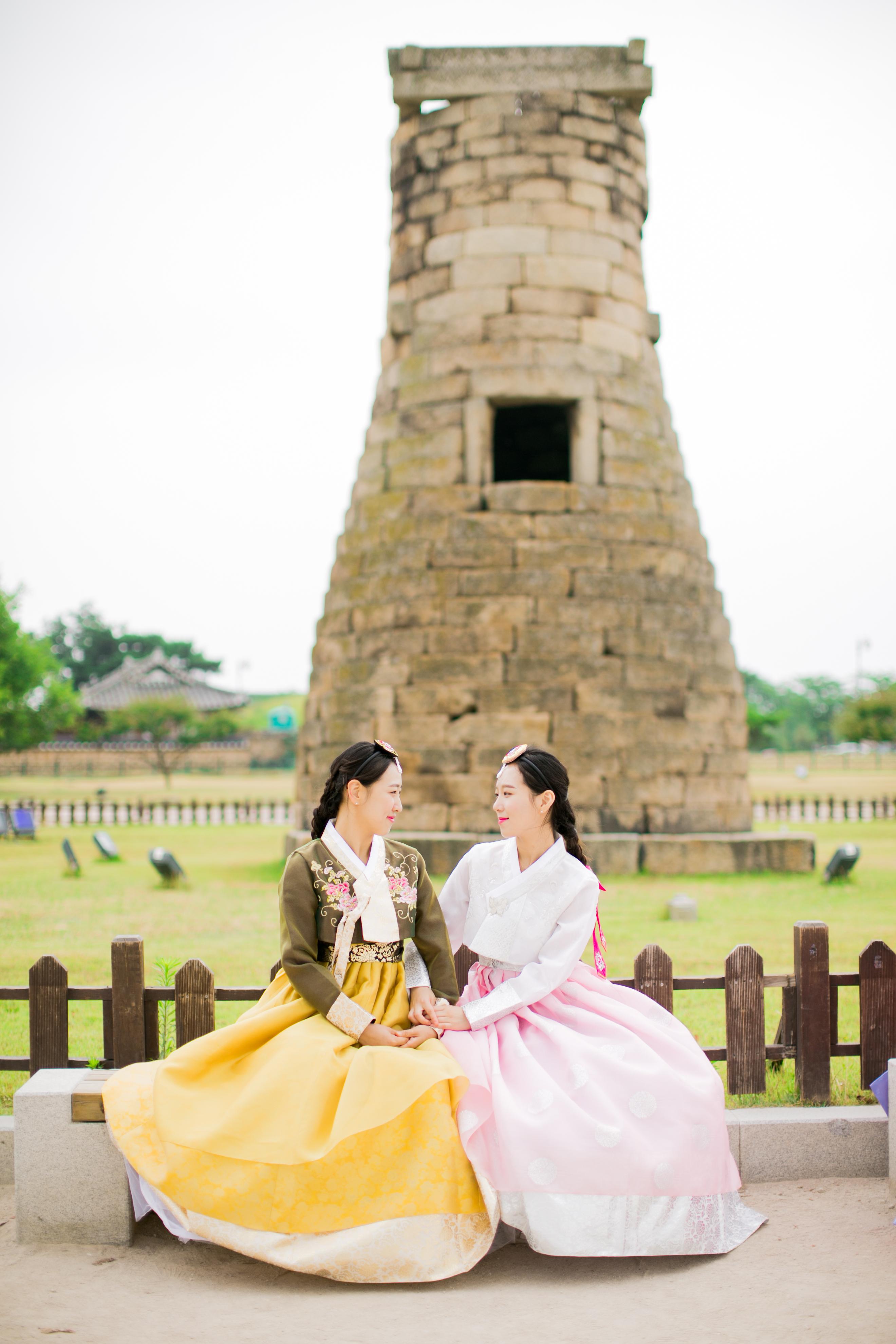 Two women in colorful hanbok (yellow and pink) sitting on a bench in front of the stone Cheomseongdae observatory, holding hands.