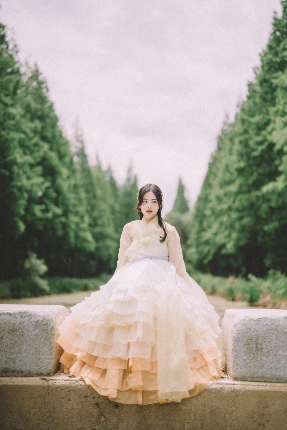 Young woman in layered pastel hanbok sitting on stone ledge framed by tall green trees in a serene outdoor setting