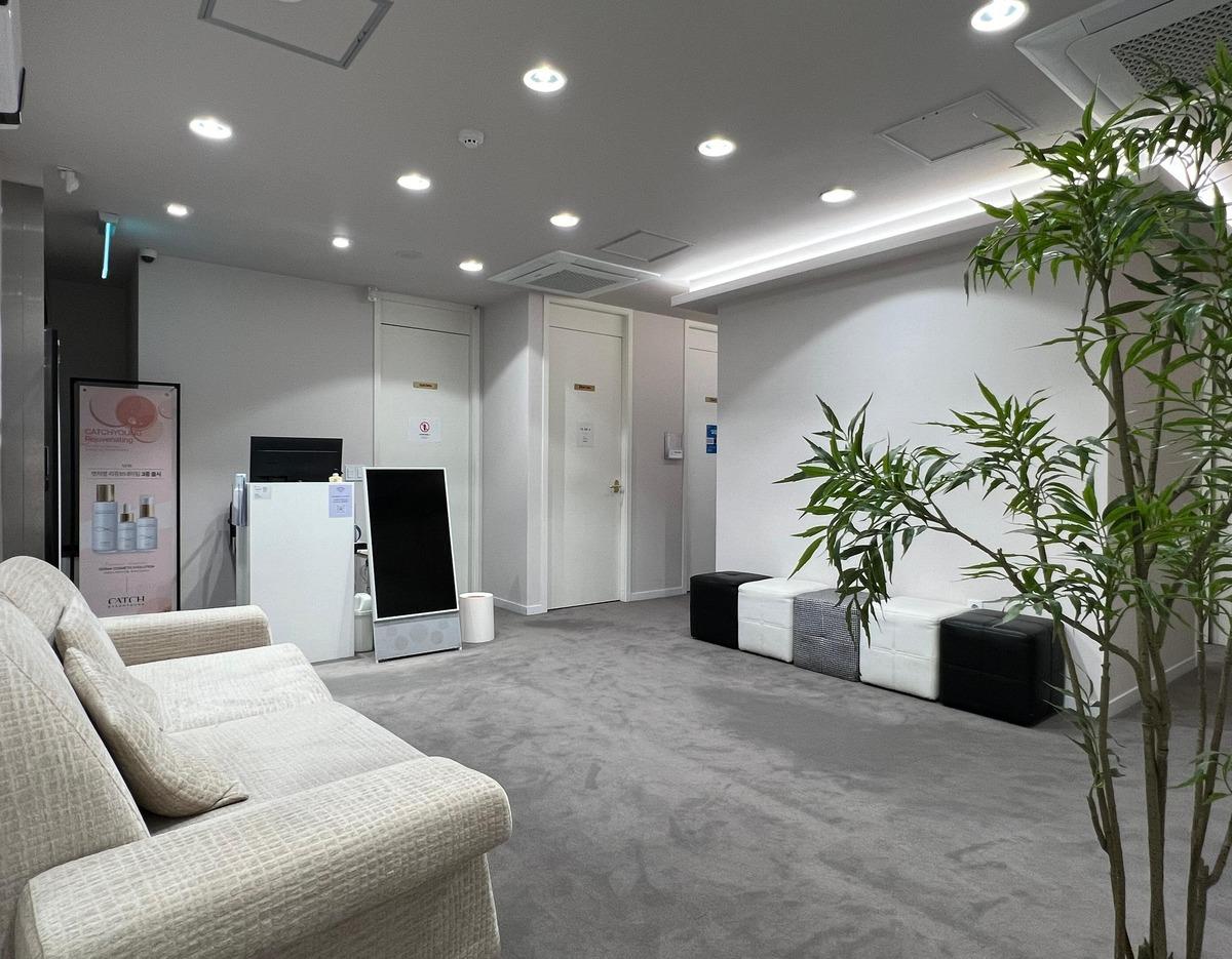Bright minimalist clinic waiting area with beige sofa, modular black-and-white ottomans, potted plant and reception kiosk.