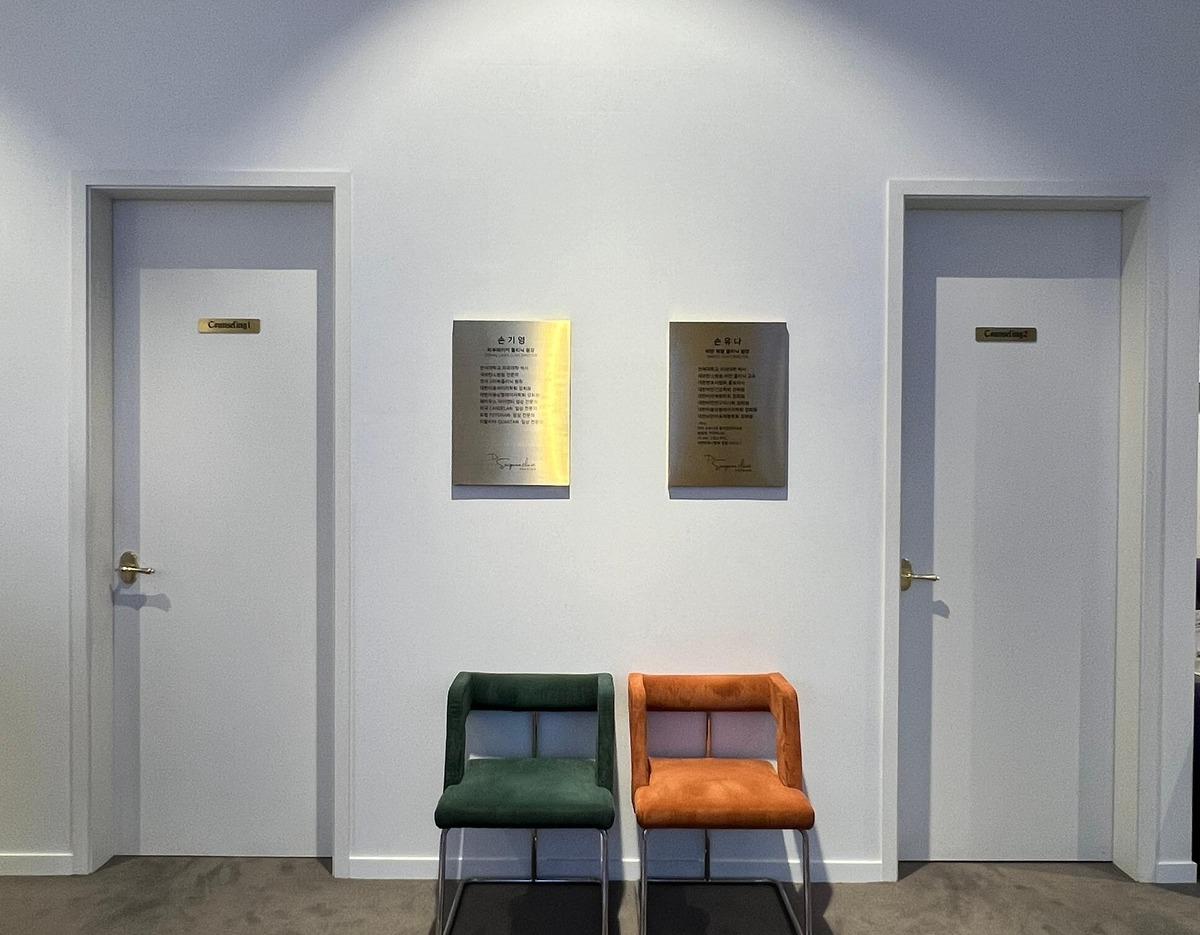 Minimal clinic waiting area with two velvet chairs (green and orange) between two white doors and brass plaques on the wall