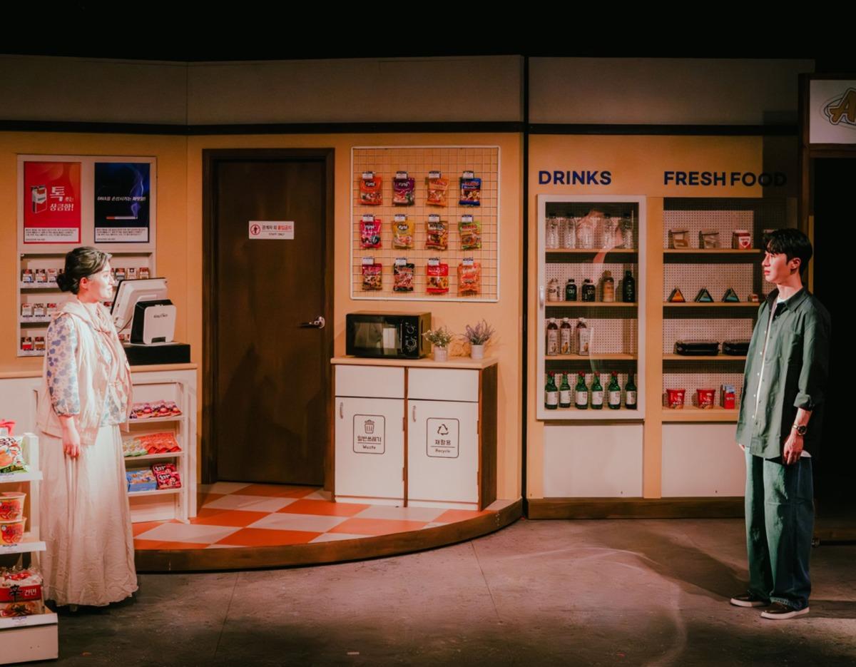 Busan Music Drama stage set of a convenience store with shelves labeled "DRINKS" and "FRESH FOOD", actress and actor facing each other.