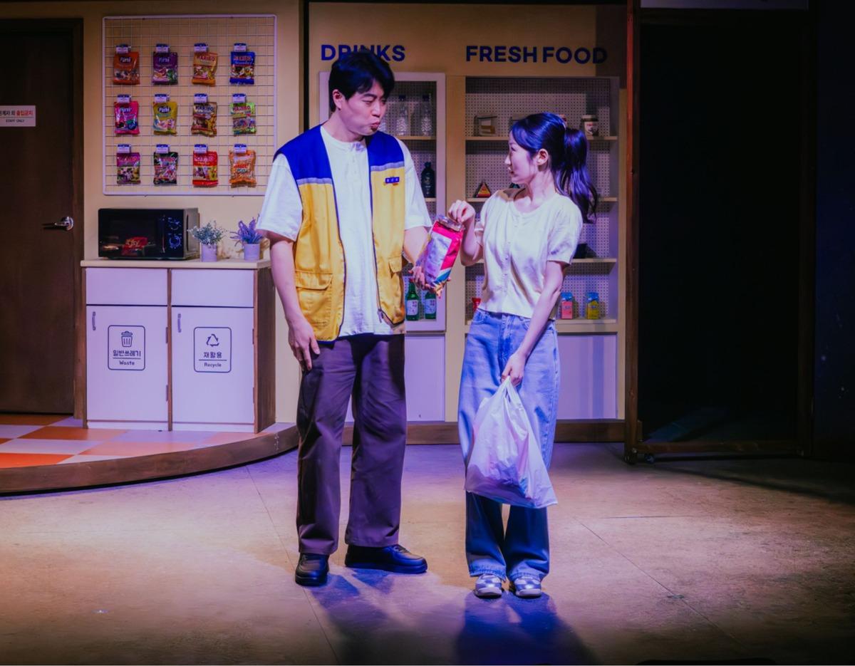 Two actors perform on a convenience store stage set; male in yellow vest hands snacks to female holding a plastic shopping bag.