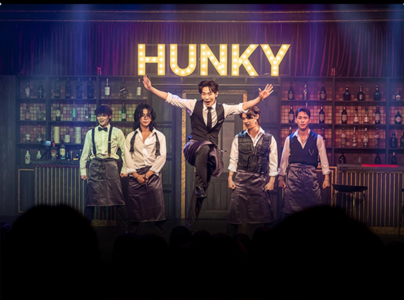 HUNKY marquee above five male performers in bar costumes, one mid-jump center stage with illuminated bottles on shelves behind