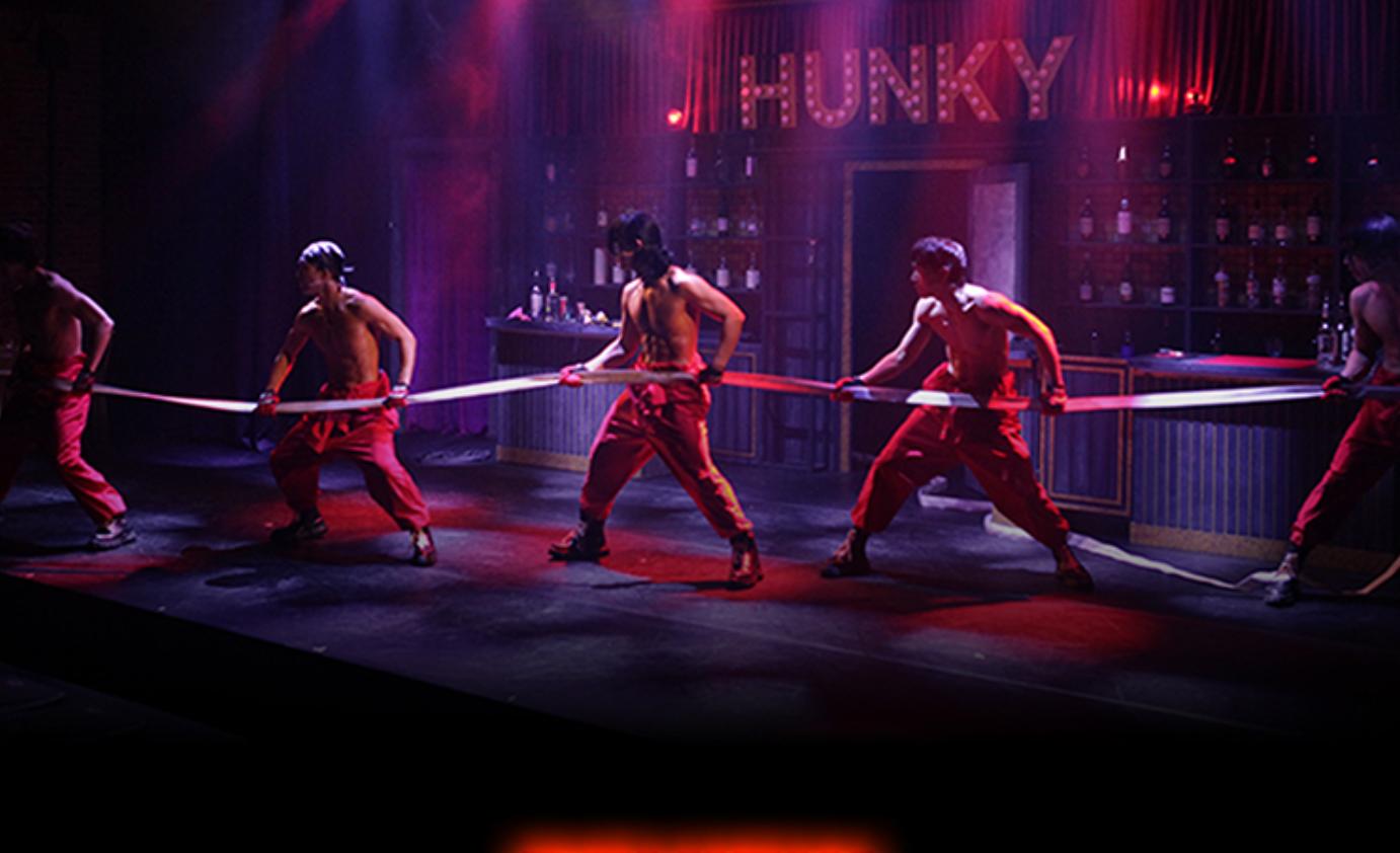HUNKY SHOW stage: five shirtless male performers in red pants pulling a long ribbon under purple-red stage lighting with bar backdrop.
