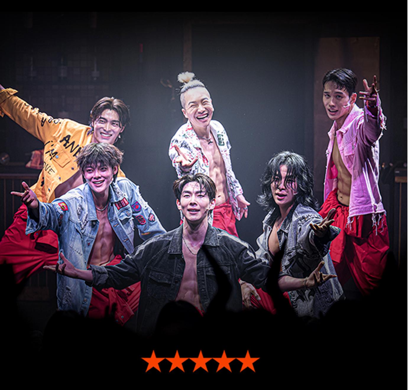 Six male performers on stage in open shirts and red pants, smiling and reaching toward audience under dramatic lighting with five stars below