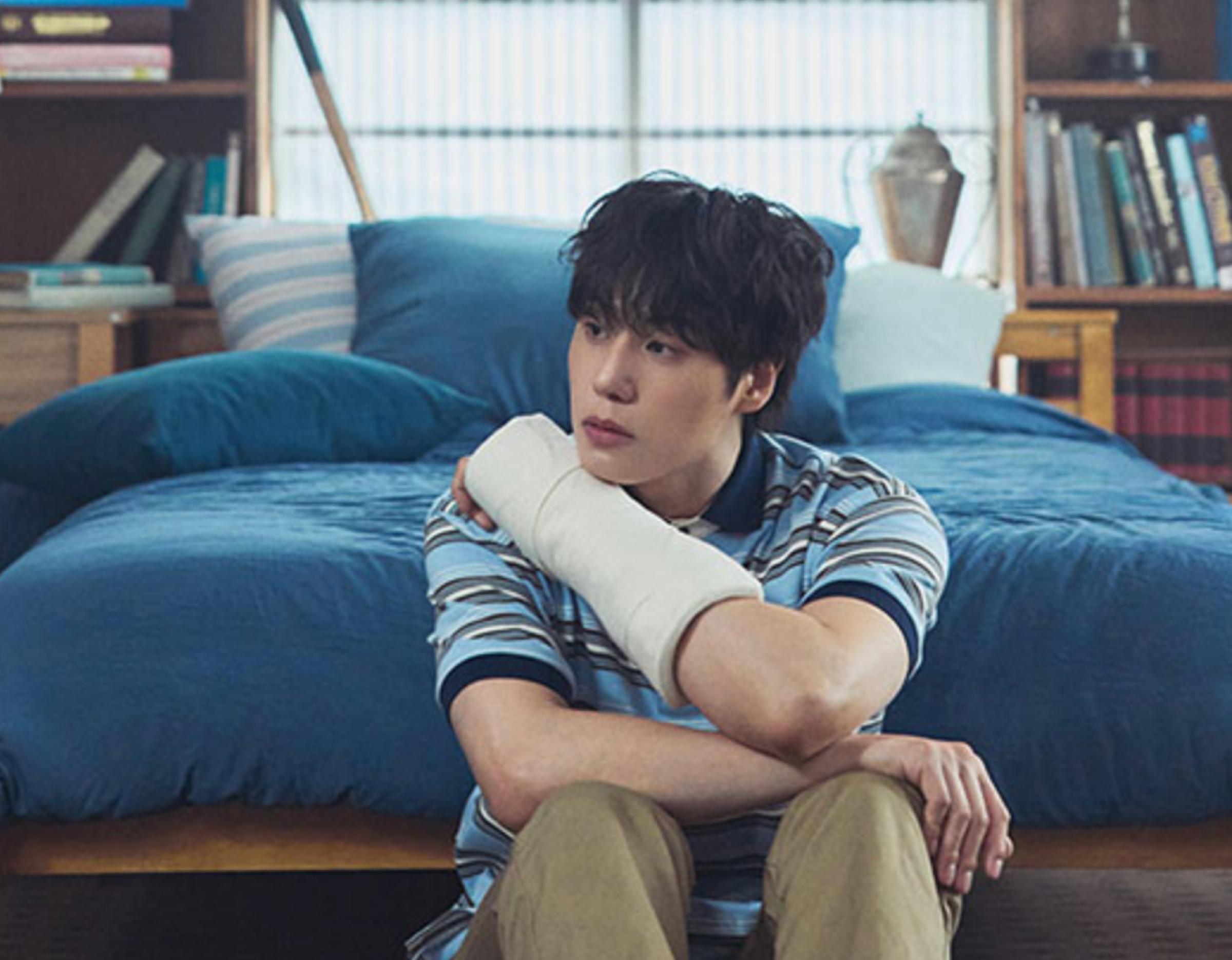 young man with a white arm cast sitting on floor in front of a blue bed, looking pensively to the side
