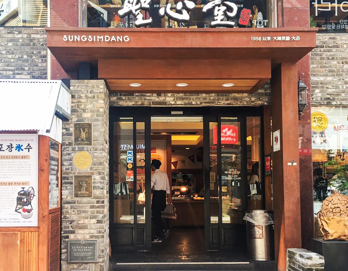 Sungsimdang bakery entrance with glass double doors, brick facade, wood awning and a person inside carrying a bag